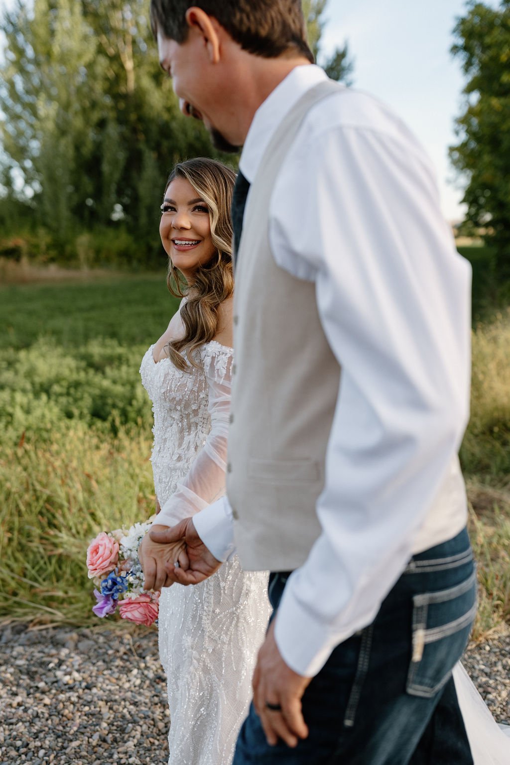 A bride smiling and looking at the groom, holding a bouquet, outside in a natural setting during daytime, with a man holding her hand.