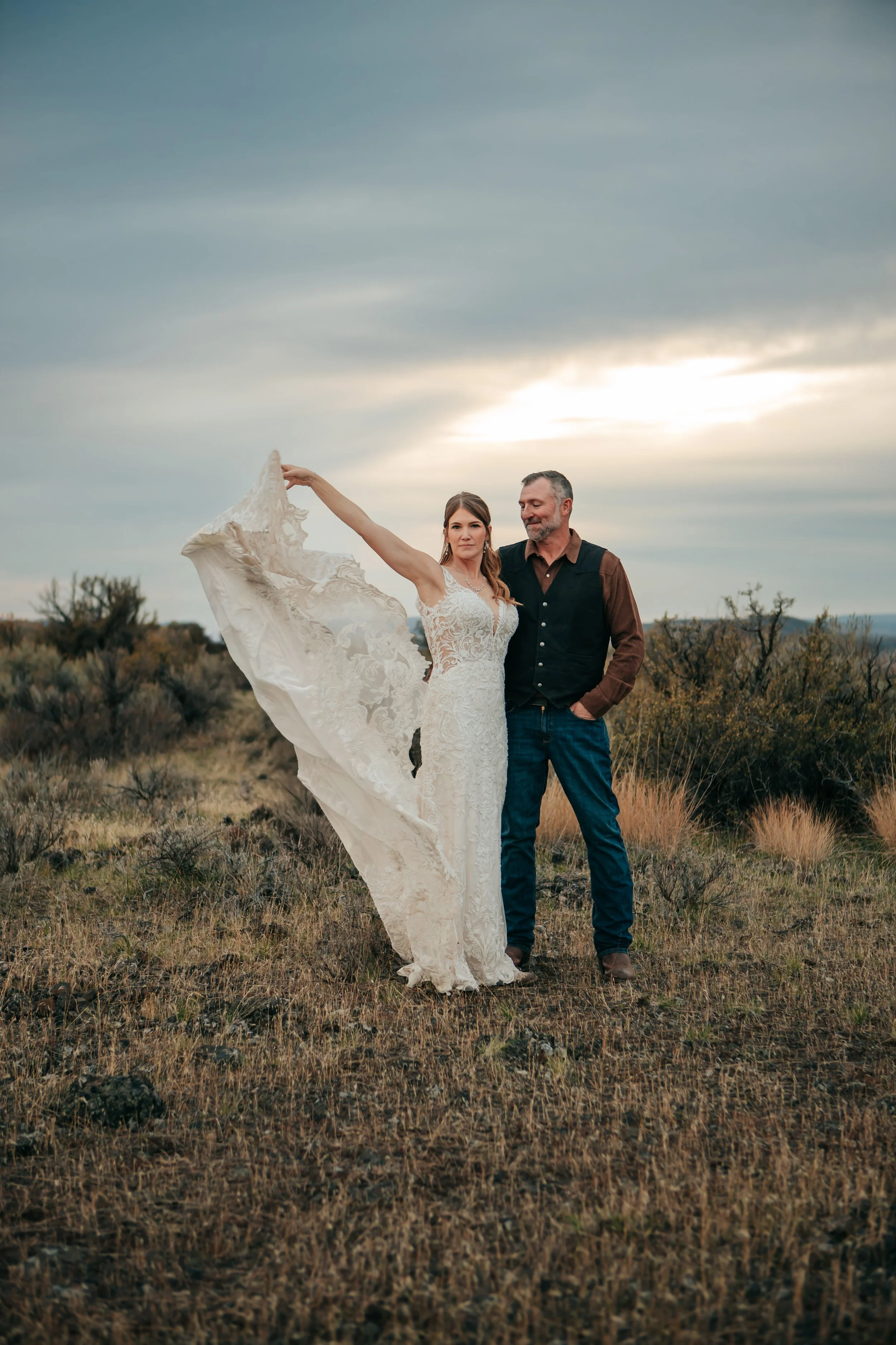 A newlywed couple standing in a grassy, rocky field at sunset. The bride wears a lace wedding dress, holding part of it up with her arm extended, and the groom stands beside her with his hand in his pocket, both smiling softly.