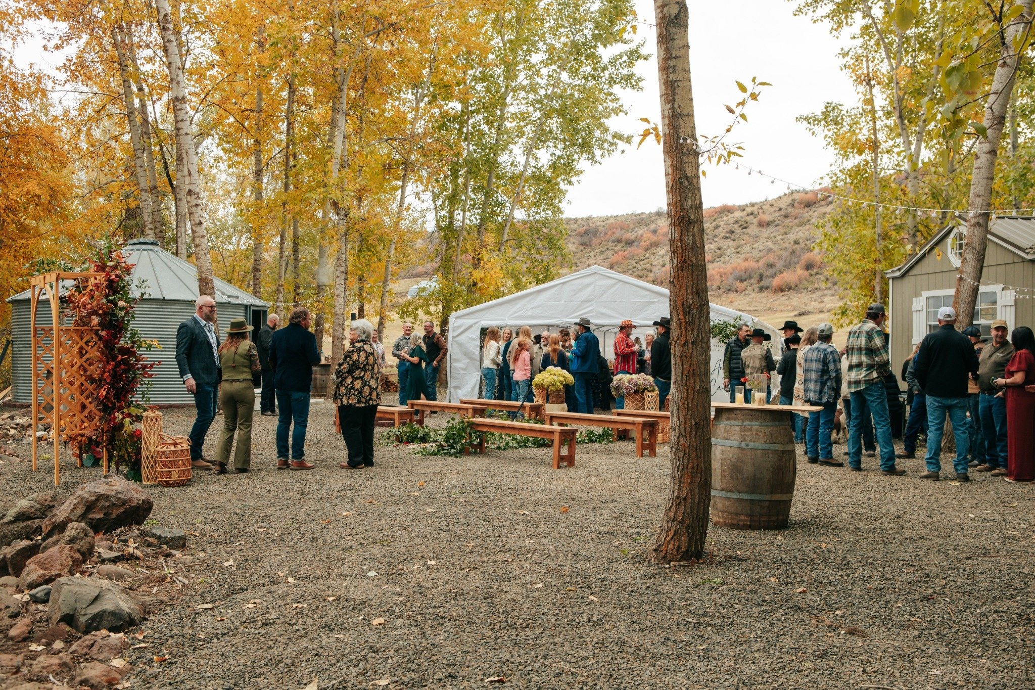 People gather outdoors at a fall event with colorful trees, white tents, and string lights in a mountainous landscape.