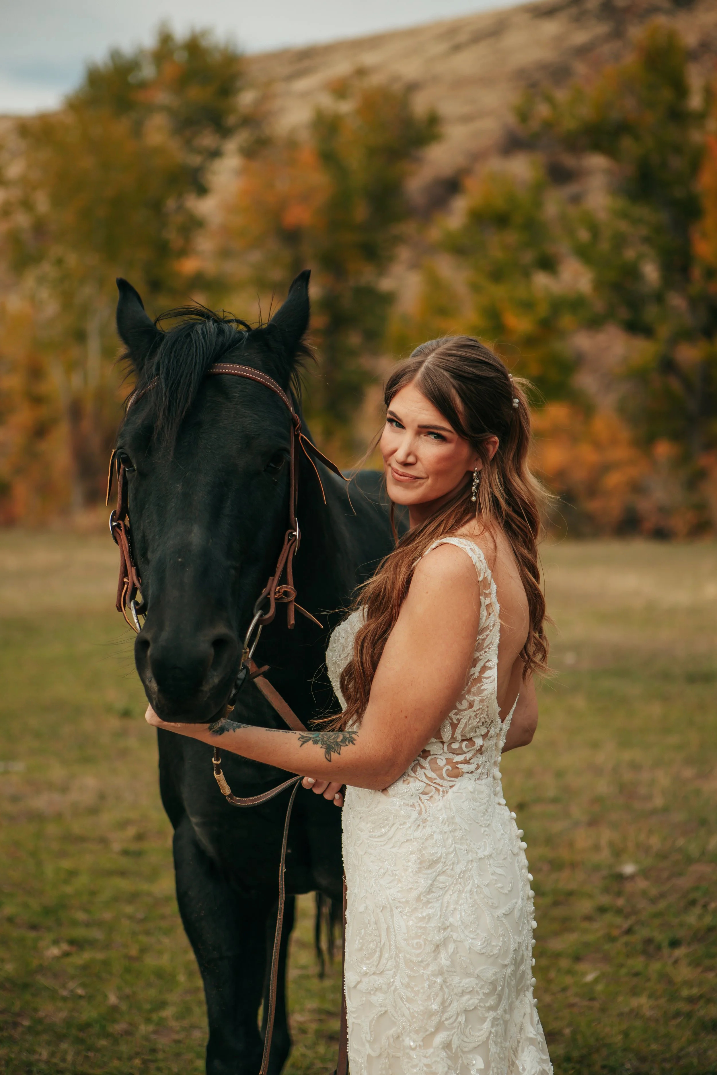 A woman in a white lace dress with long brown hair, earrings, and a tattoo on her arm, standing outdoors with a black horse, with autumn trees and a hill in the background.