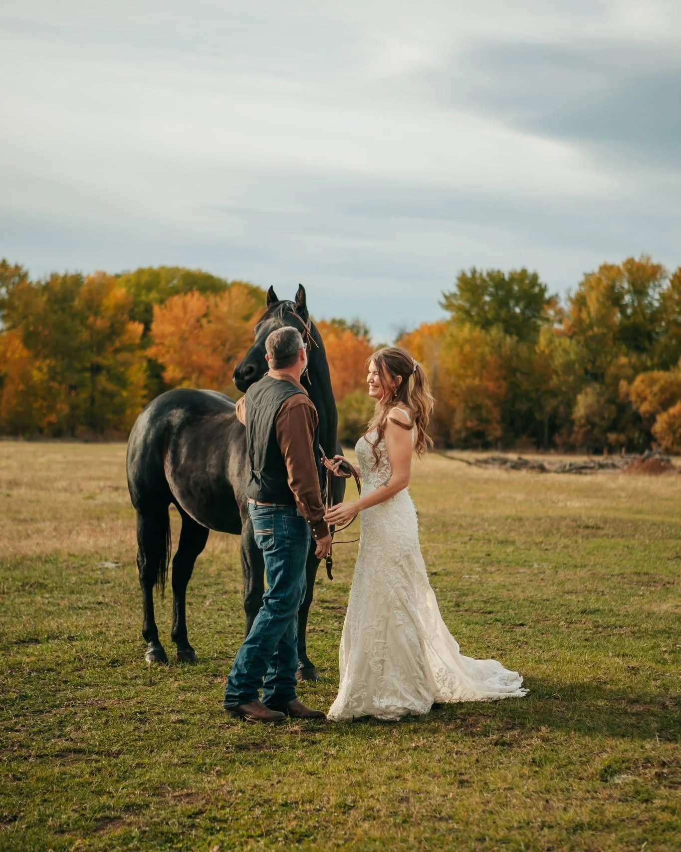 A man and woman in wedding attire standing with a black horse in an open field, with autumn trees in the background.