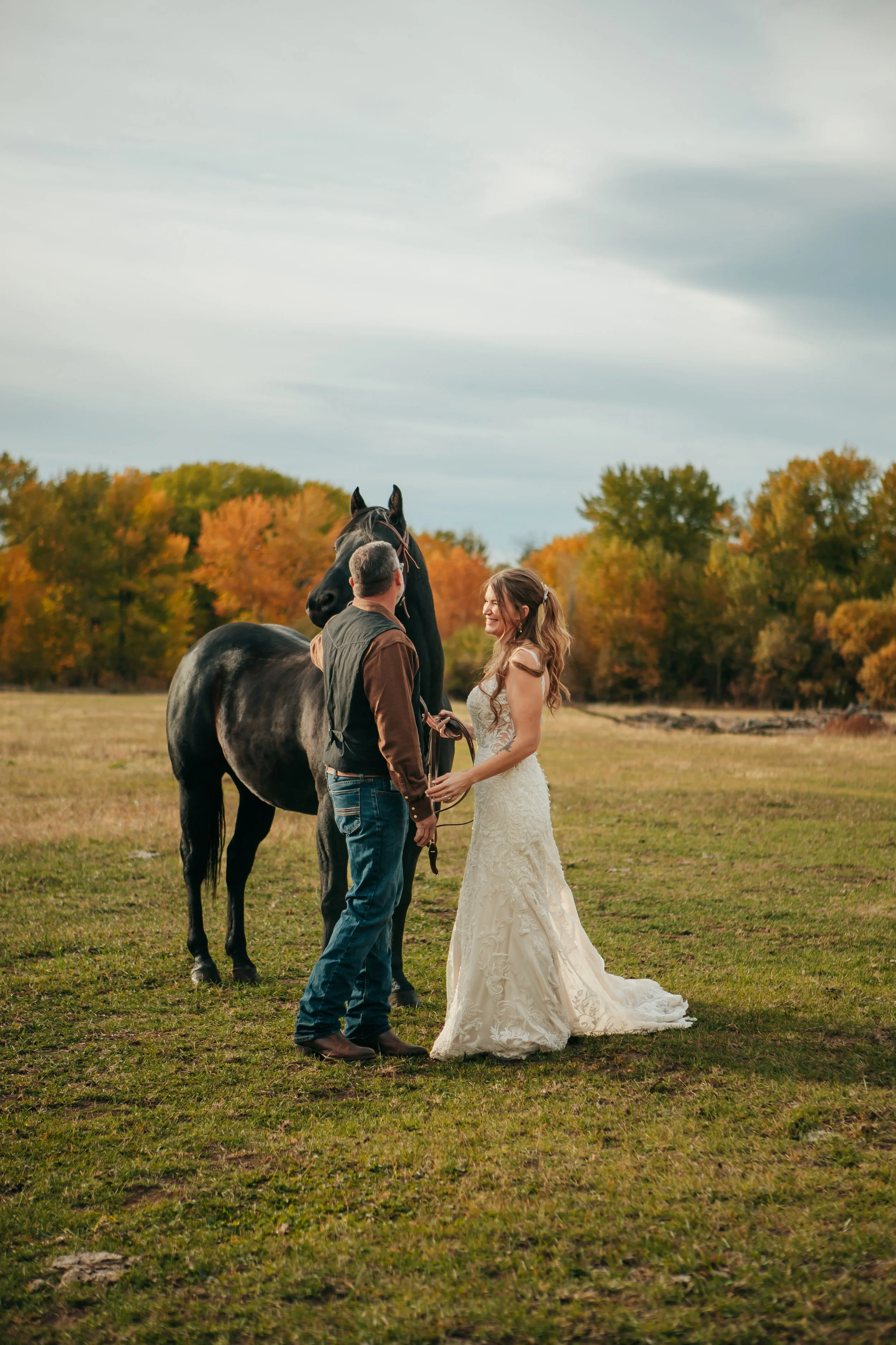A bride and groom at Riverwild holding hands and smiling at each other on a grassy field during sunset, with a man holding a black horse's bridle. Autumn trees with orange and green leaves are in the background, and the sky is partly cloudy.