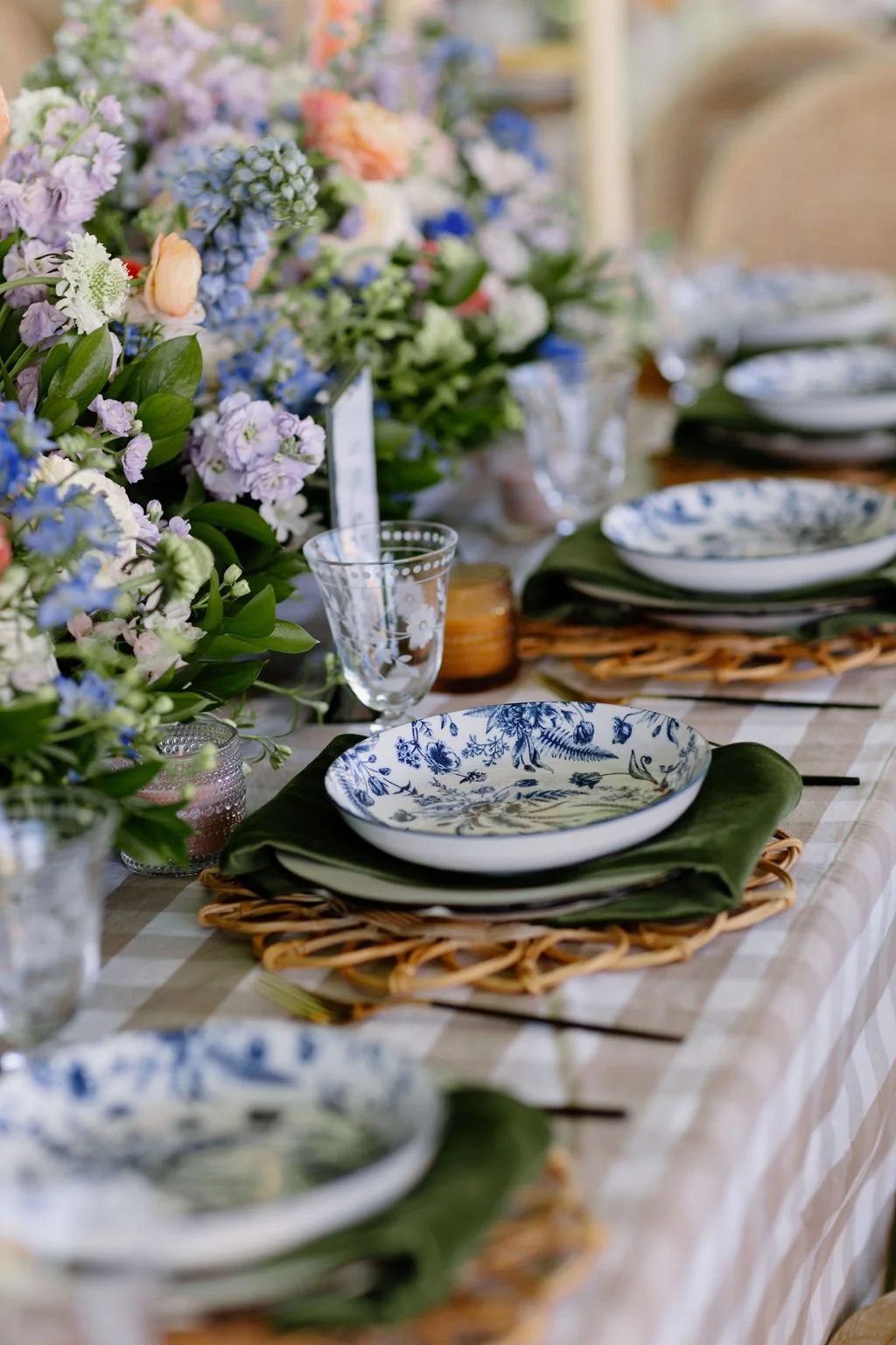 A long dining table decorated with floral centerpieces, plates with blue floral patterns, wine glasses, and dark green cloth napkins on woven placemats, set for a meal.
