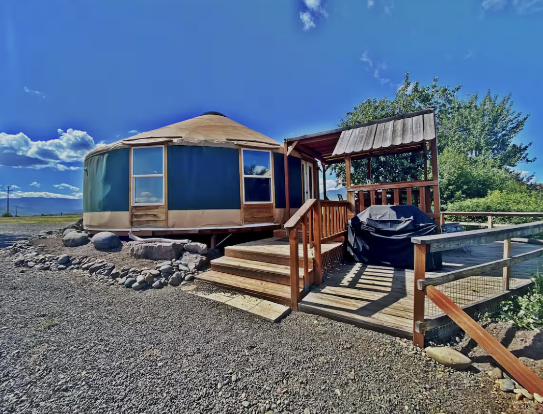A yurt with a wooden deck and stairs, with a grill covered in a black cover on the deck, outdoors on a clear day with blue sky and trees in the background.