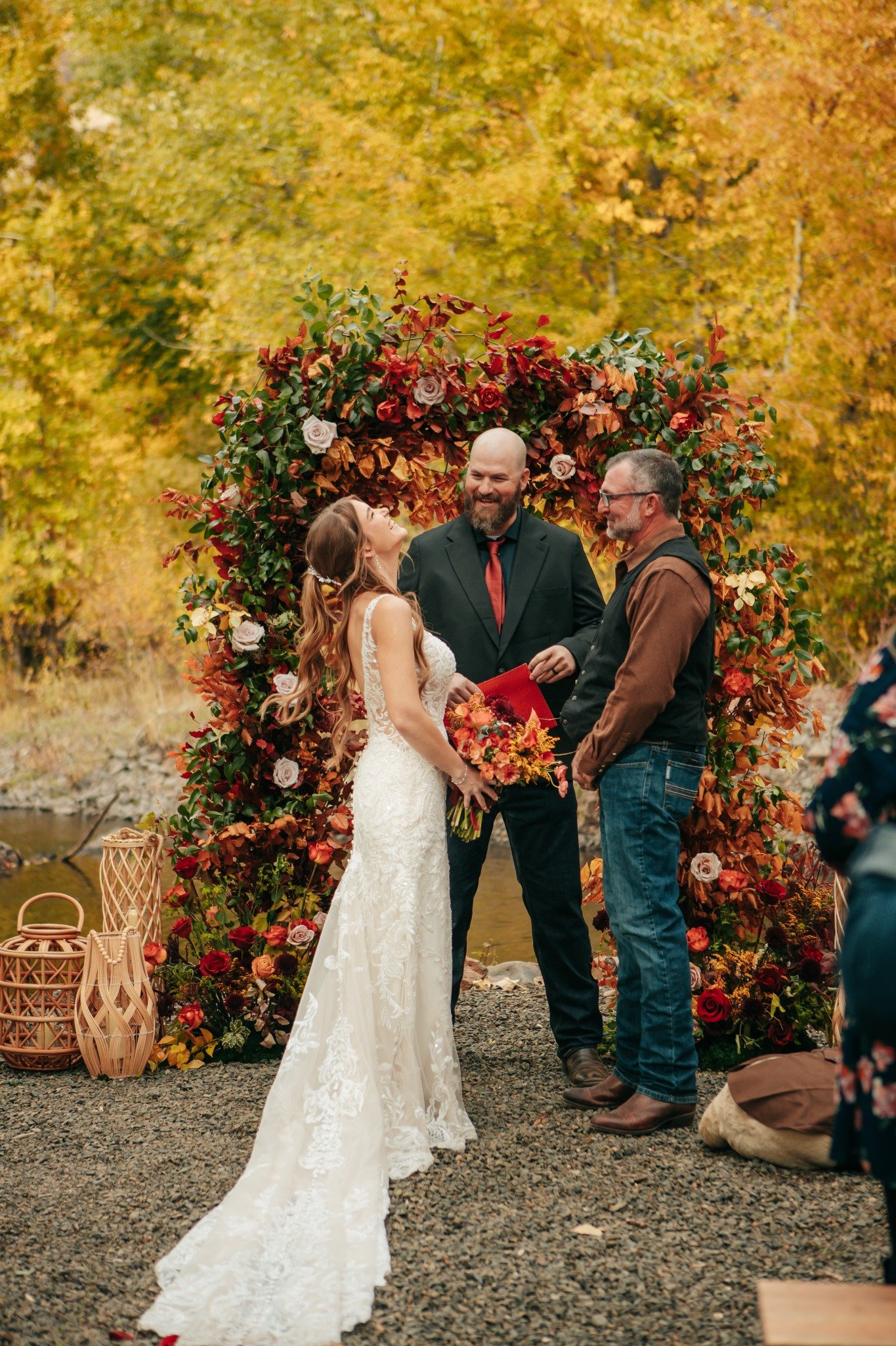 A wedding ceremony outdoors in autumn with a couple exchanging vows, surrounded by a floral arch, onlookers, and autumn trees with orange leaves.