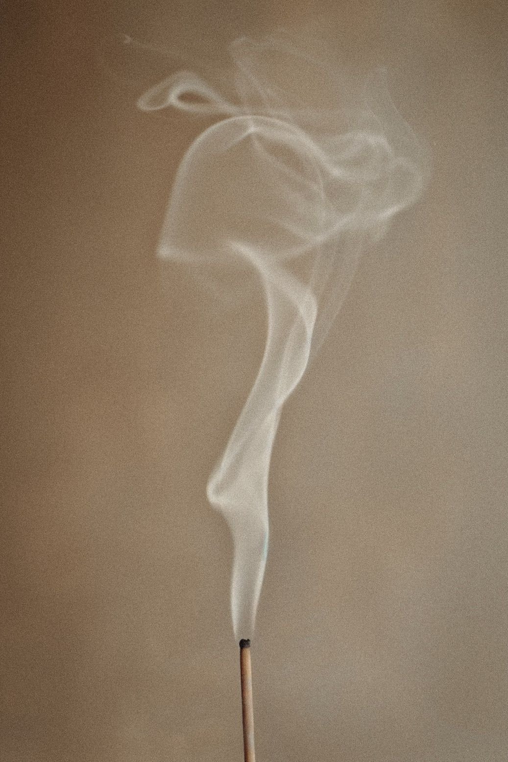 A lit incense stick releasing a trail of smoke in various swirling patterns against a beige background.