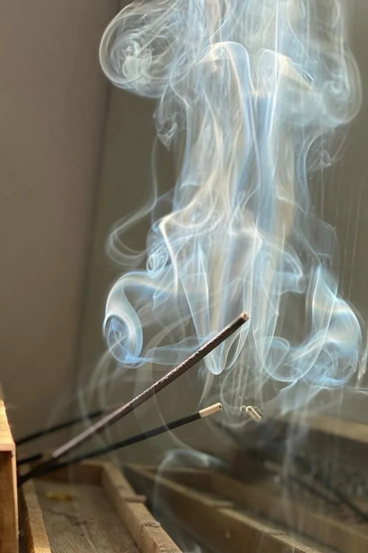 Close-up of a lit incense stick emitting swirling smoke in a room with wooden structures.