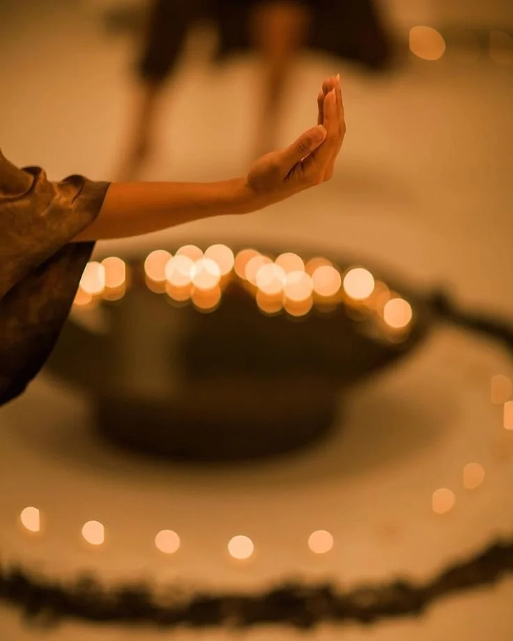A person practicing meditation or prayer, with their hand raised and fingers together, in a dimly lit room with candles or fairy lights creating a warm, peaceful ambiance.