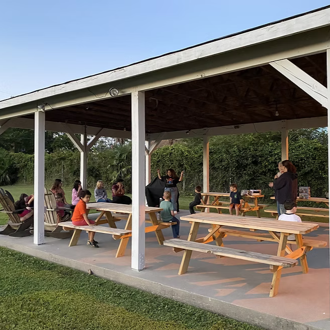 Children and adults gathered under a pavilion with picnic tables, some sitting and others standing, with a woman hosting a presentation or activity in front of a grassy area and trees.