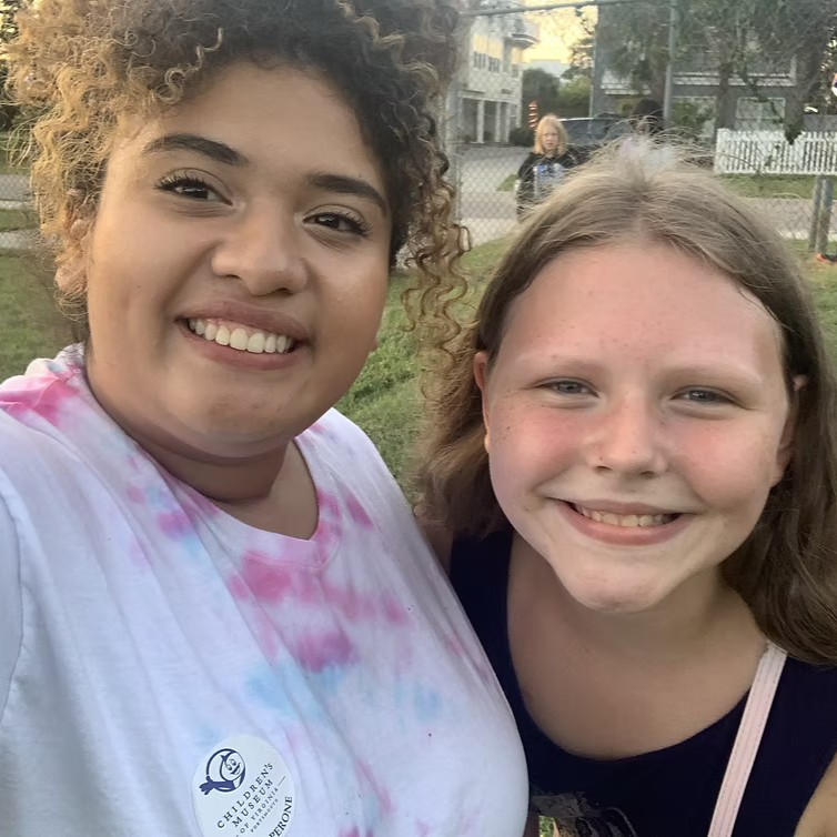 Two young girls smiling for a selfie outdoors, with a fence and trees in the background.