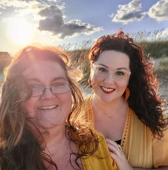 Two women smiling outdoors on a beach at sunset, one with glasses and long wavy hair, the other with dark curly hair and glasses, both wearing yellow tops.
