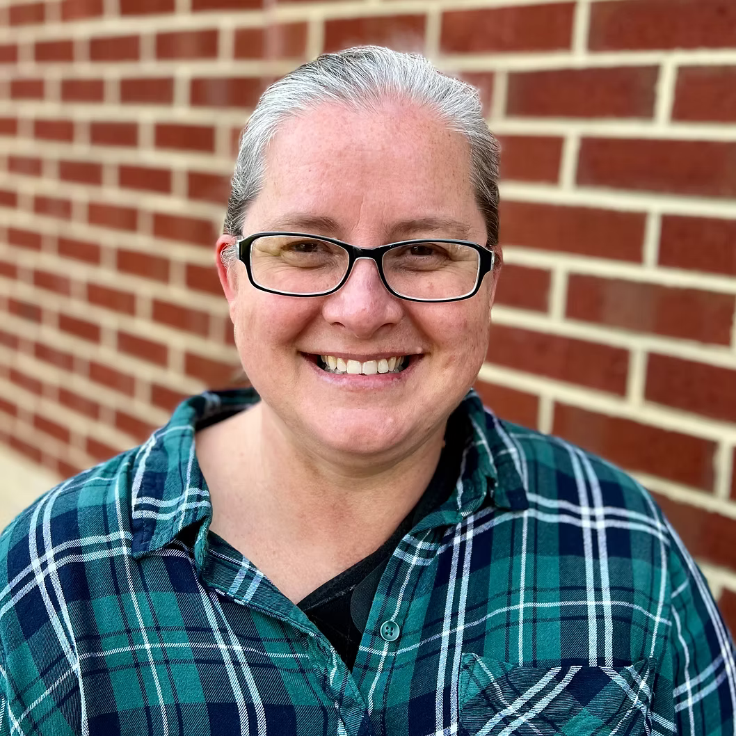 A smiling person with glasses and short gray hair wearing a green and blue plaid shirt standing in front of a red brick wall.