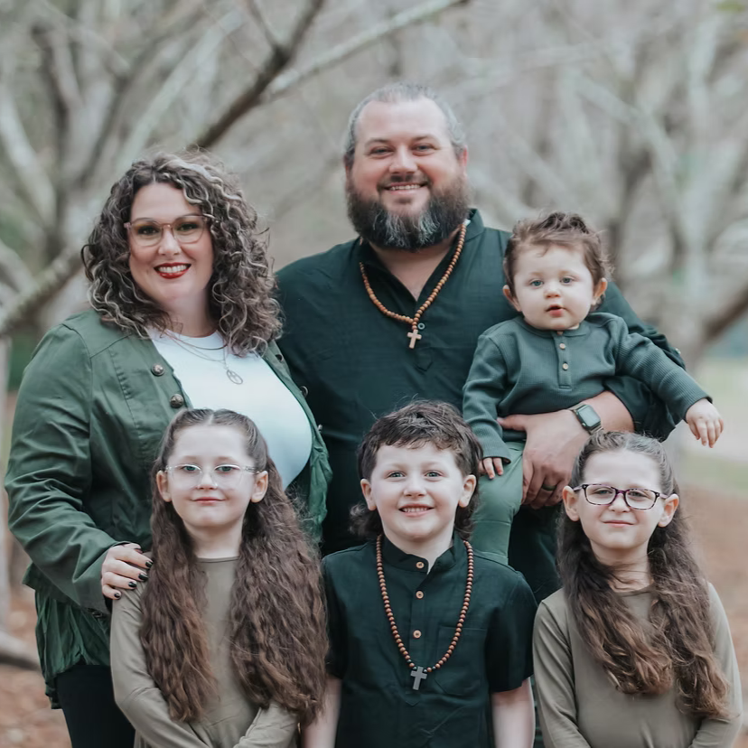 Family photo outdoors featuring a woman, a man, three girls, and a toddler boy, all smiling, with trees in the background.