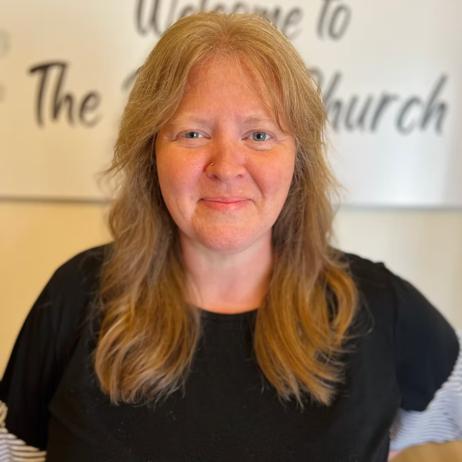 A woman with light brown hair and blue eyes, smiling, standing in front of a sign that says "Welcome to The Church."