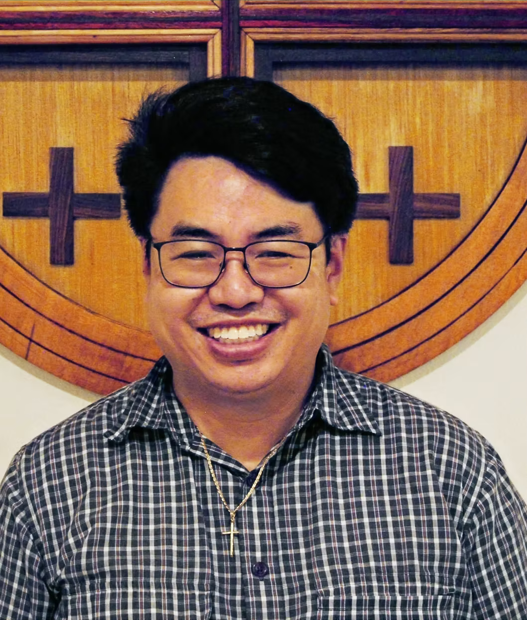 A smiling man wearing glasses and a checkered shirt, standing in front of a wooden wall with a large circular emblem featuring crosses.