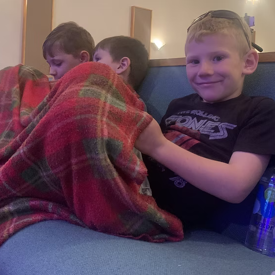 Four boys sitting on a blue couch; three covered with a red plaid blanket, one smiling boy with glasses and a Black Sabbath t-shirt visible.