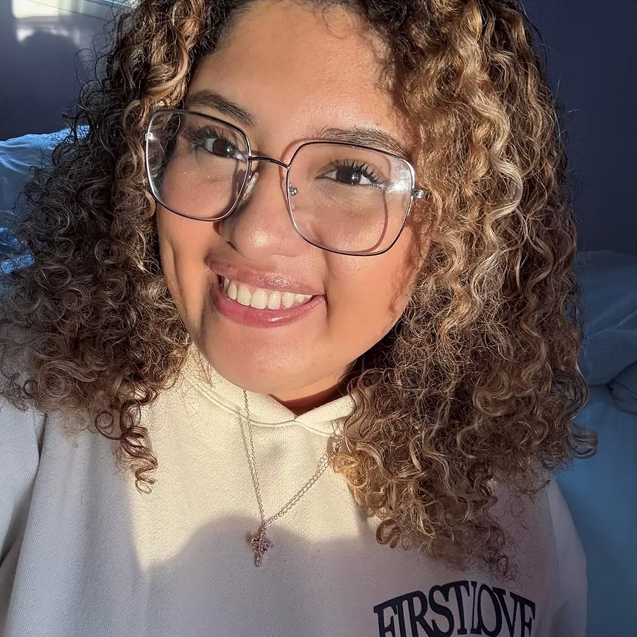 A woman with curly hair, glasses, and a bright smile. She is wearing a cream-colored shirt with a necklace and is in a well-lit room.