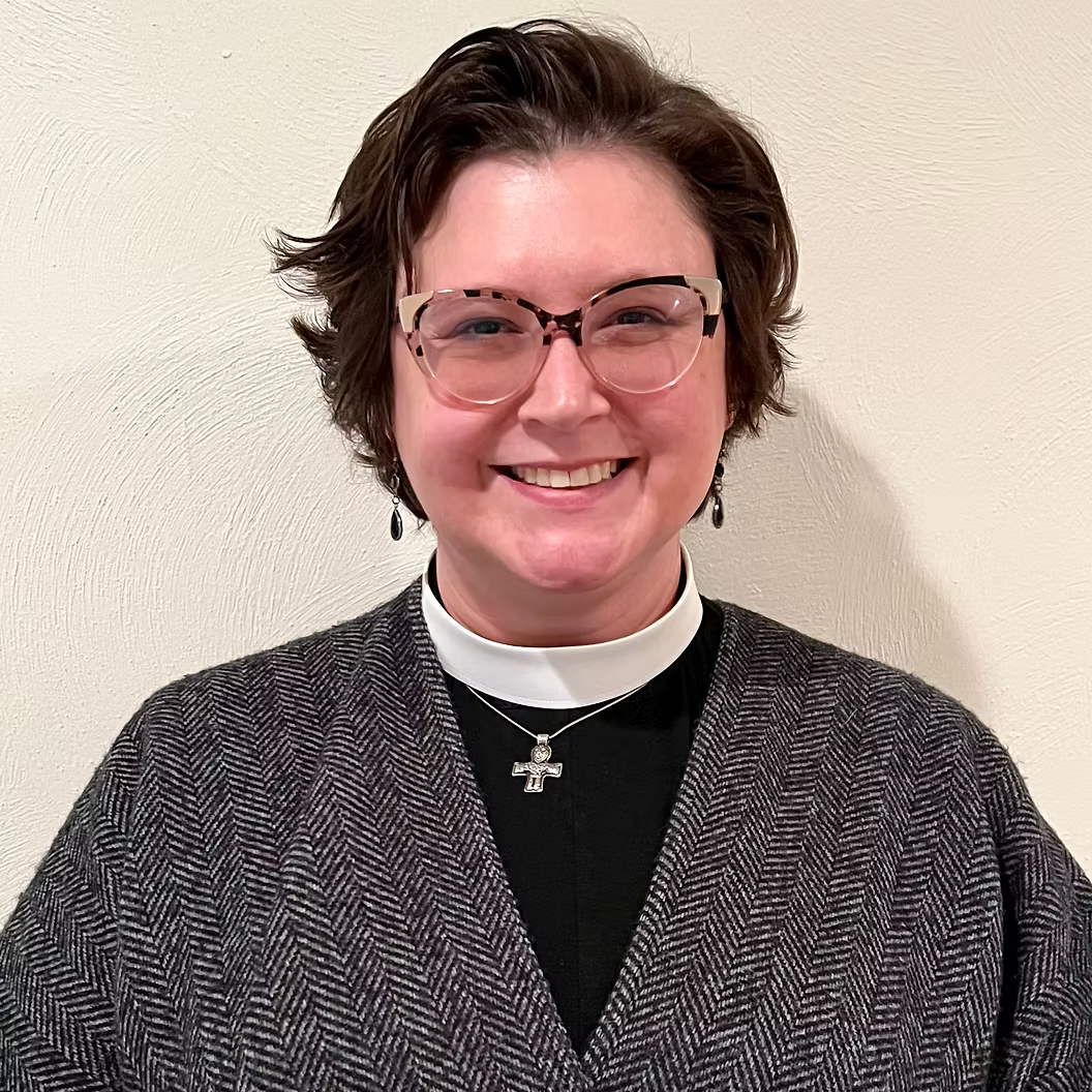 A woman with short brown hair, wearing glasses, a black top, a gray patterned cardigan, and a clerical collar with a cross necklace, smiling in front of a plain light-colored wall.