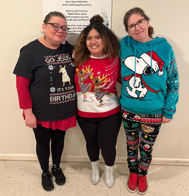 Three women standing together at a Christmas celebration wearing holiday sweaters. The woman on the left is wearing a black sweater with a llama design and the words "It’s Your Birthday." The woman in the middle is wearing a red sweater with a reindeer and reindeer antlers. The woman on the right is wearing a bright blue sweater with Snoopy wearing a Santa hat. They are smiling and standing against a white wall.
