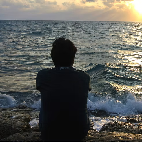 Person sitting on rocks by the ocean at sunset, looking at the water.