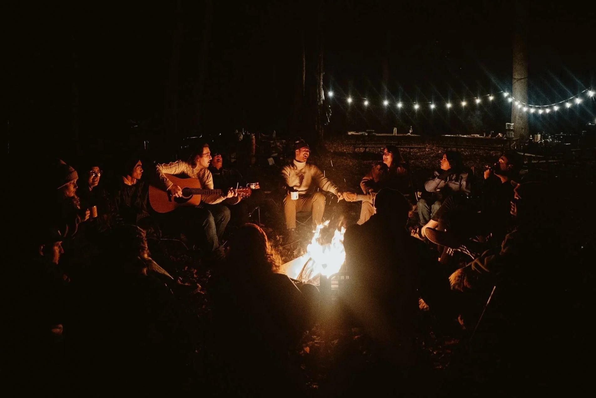 Group of people sitting around a campfire outdoors at night, some playing guitar and others talking and laughing under string lights.