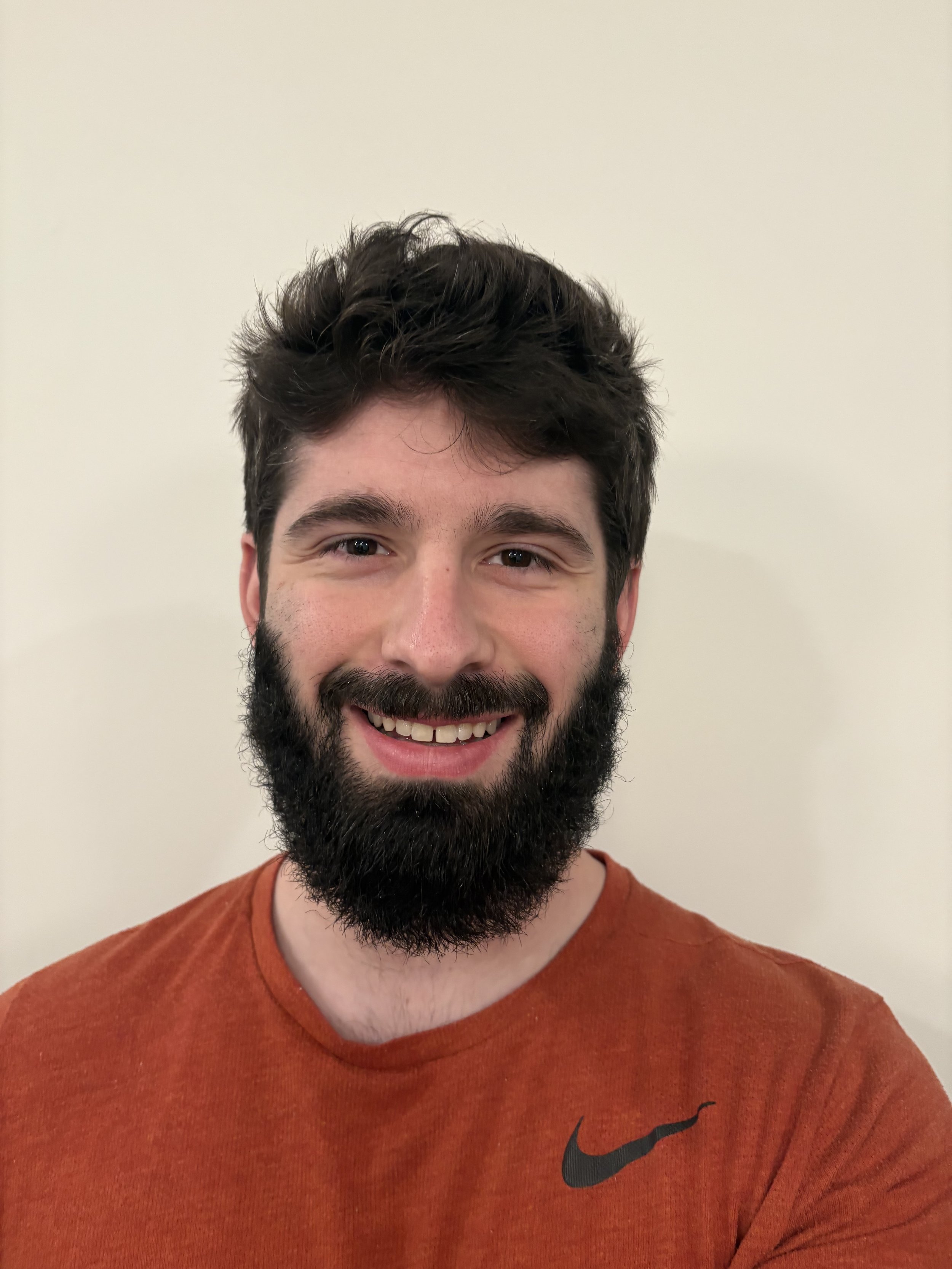 A smiling man with dark hair, a full beard, and mustache, wearing a reddish-orange shirt with a black Nike logo, standing against a plain off-white wall.