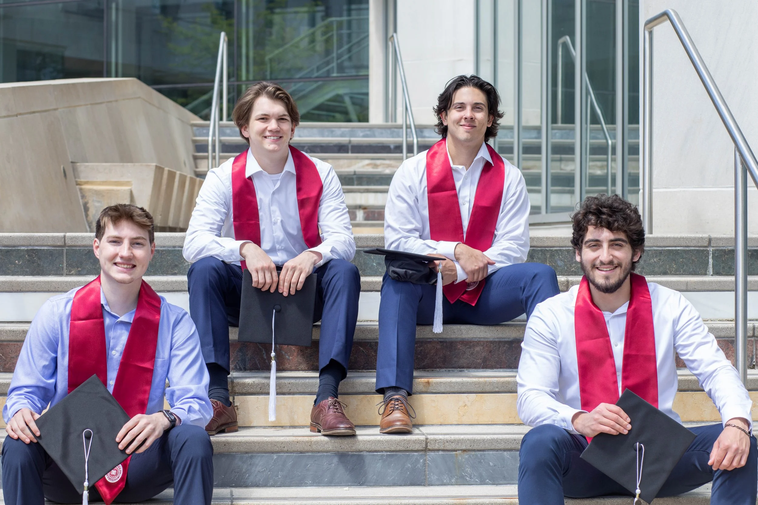 Group of five young men in formal attire with red graduation stoles sitting on stairs outside a building, smiling and holding their graduation caps.