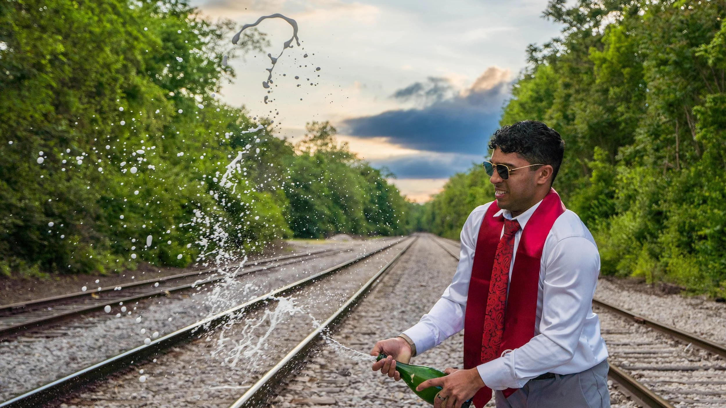 A man in formal attire, wearing sunglasses and a red scarf, is celebrating near train tracks while opening a champagne bottle, with champagne spraying into the air against a backdrop of green trees and a cloudy sky.