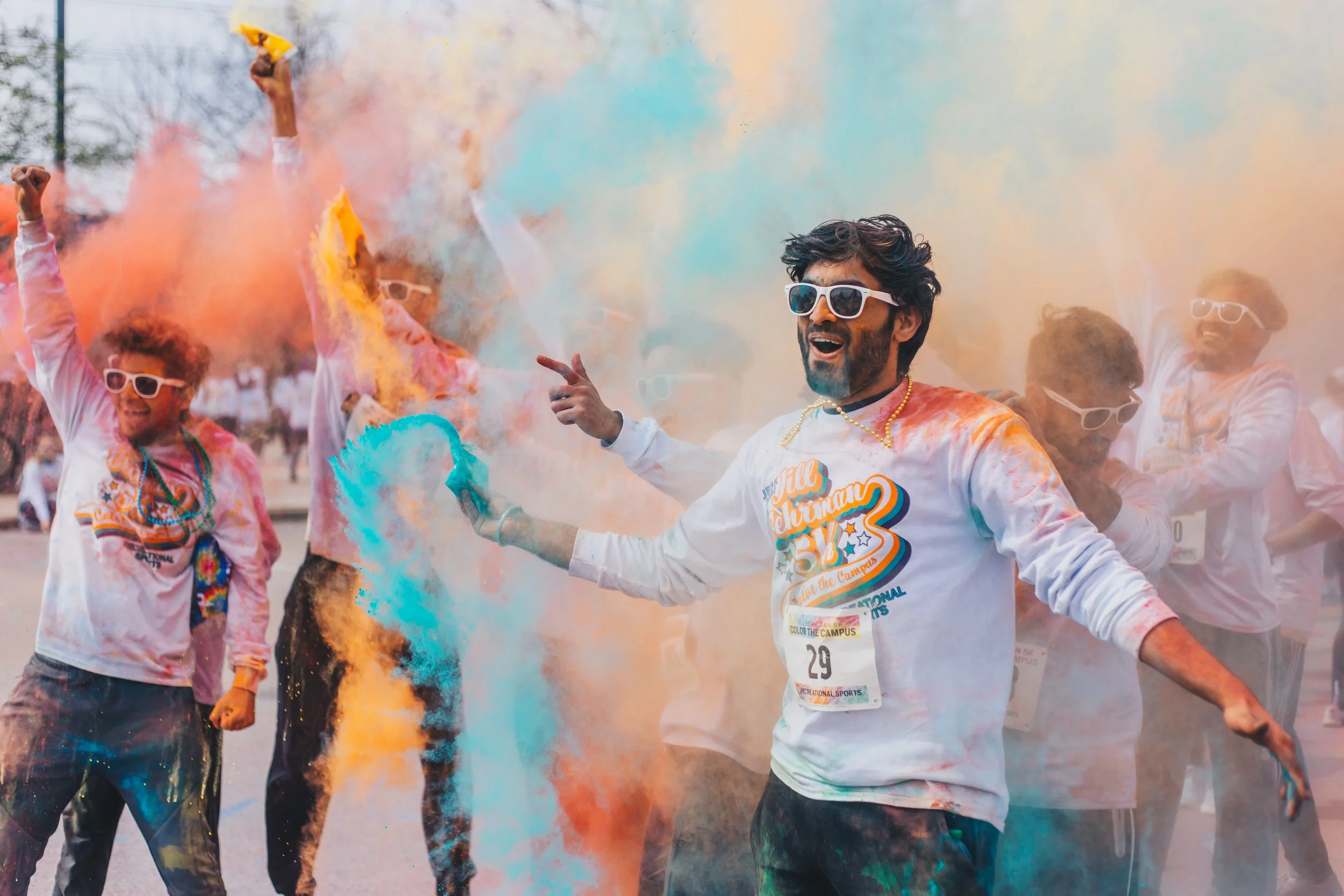 People celebrating Holi with colorful powder and smoke, wearing sunglasses and white shirts.