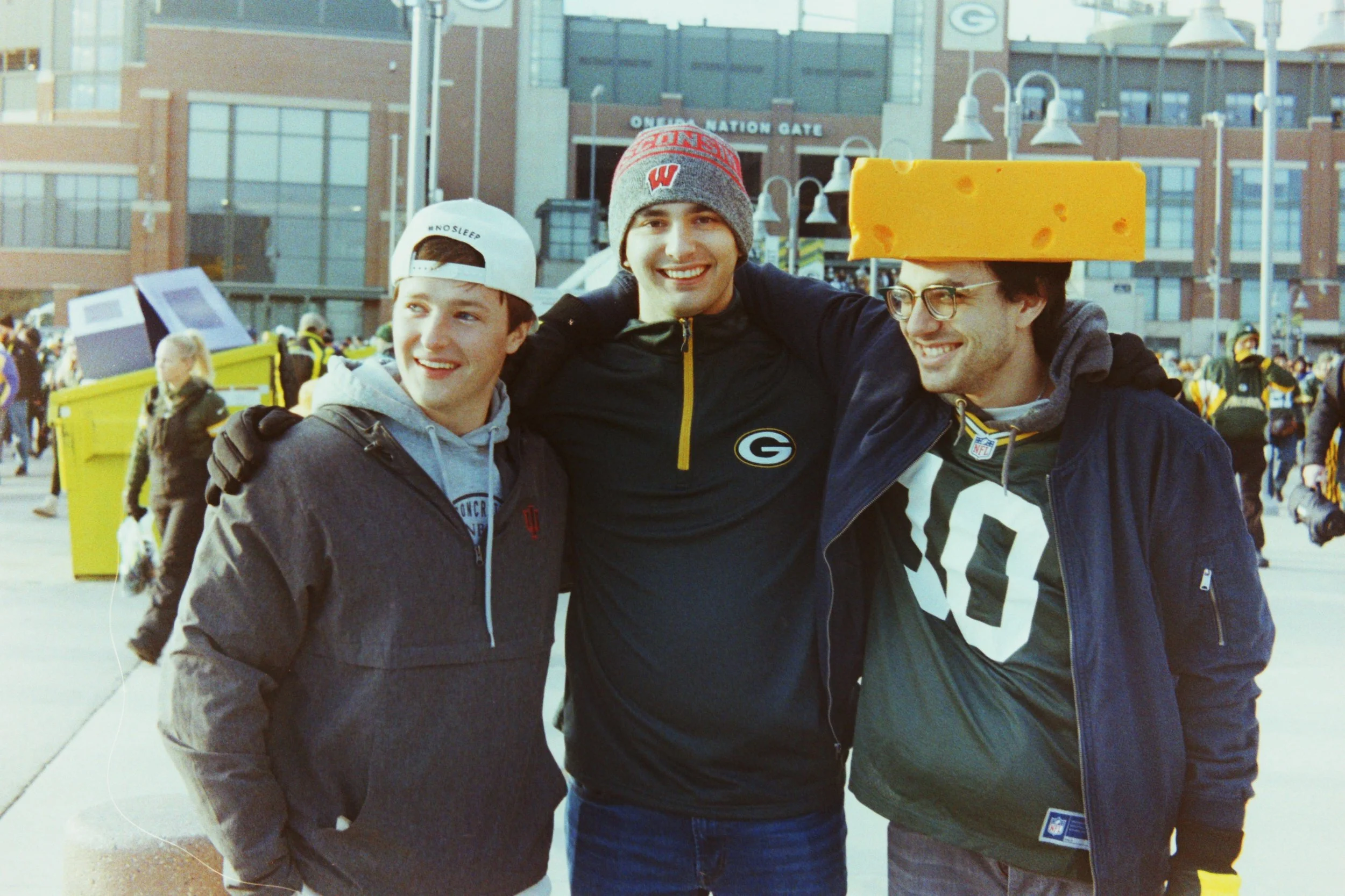 Three young men smiling with arms around each other outside, one wearing a cheese-shaped hat, in front of a crowd and a stadium entrance.