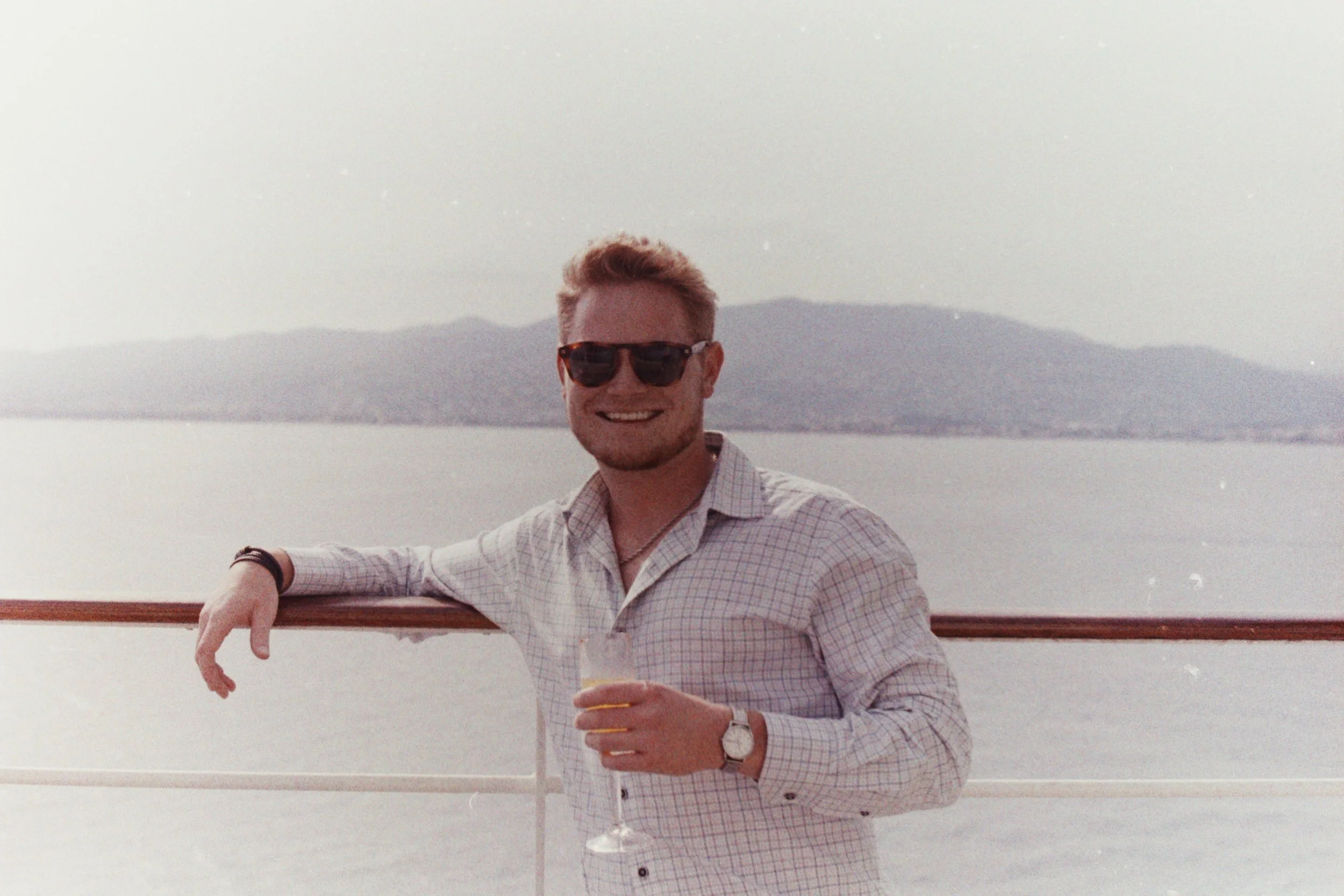 A smiling man with light hair, wearing sunglasses, a watch, and a plaid button-up shirt, stands in front of a body of water with mountains in the distance.
