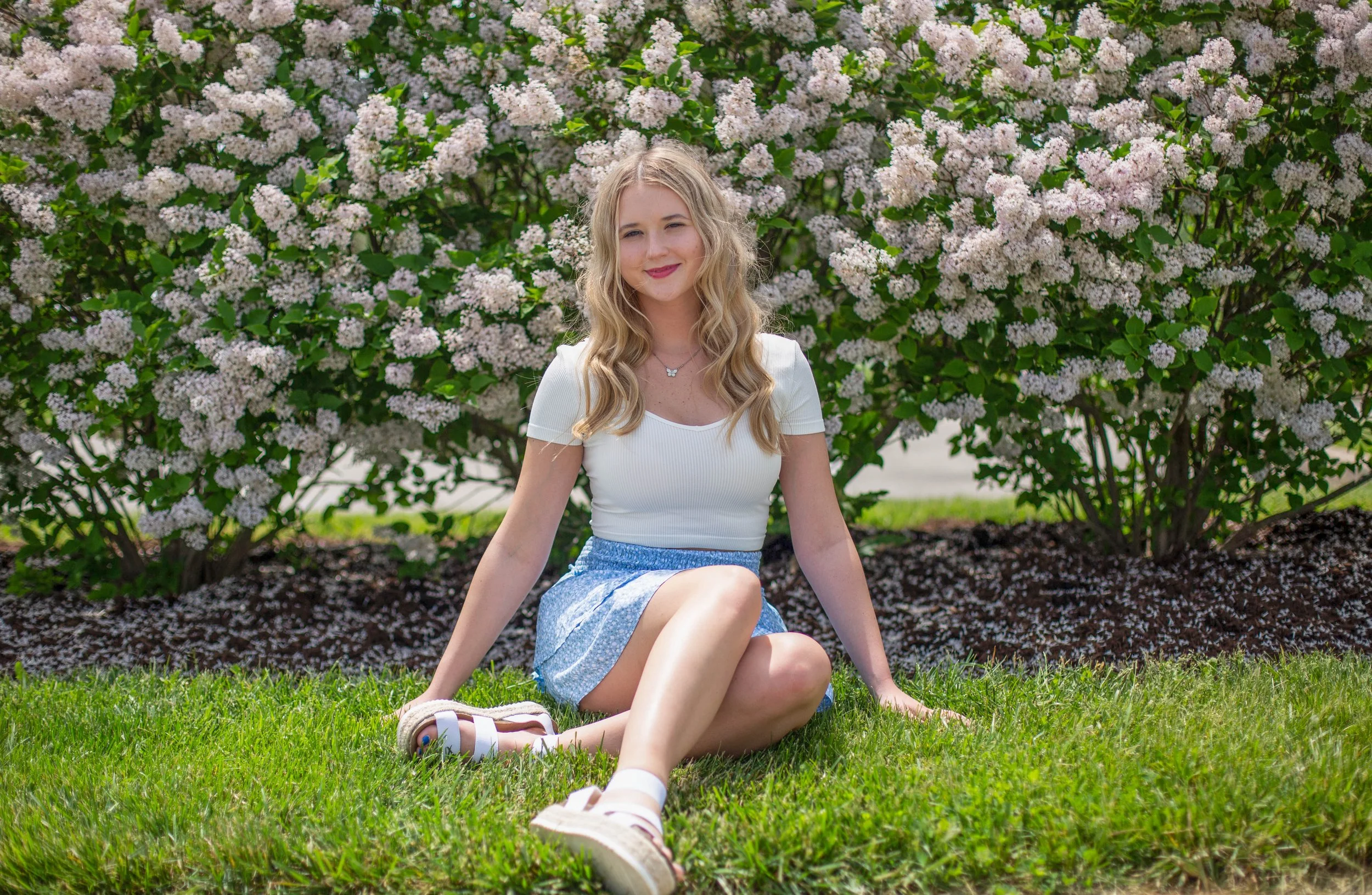 A young woman with wavy blonde hair sitting on grass in front of a large flowering shrub with white and pink blossoms, smiling at the camera.