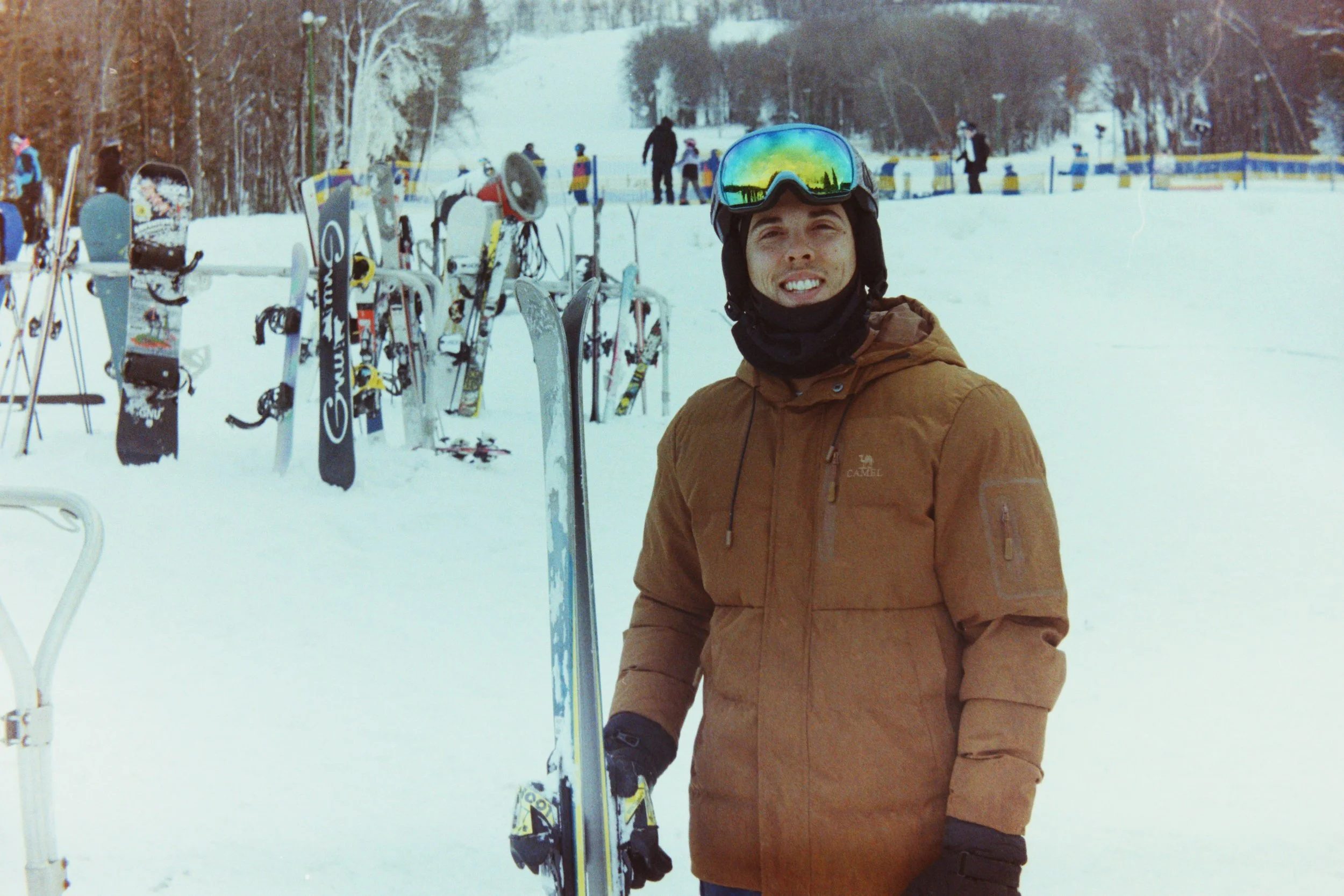 A man wearing a brown jacket, ski goggles, and a black face covering, standing in a snowy ski resort with skis and ski equipment visible, and others in the background.