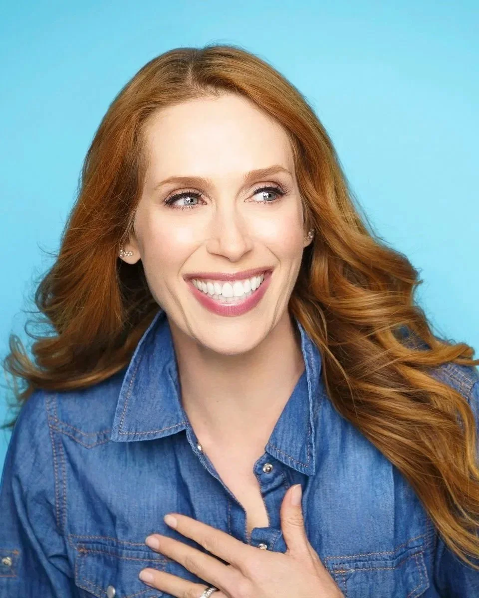 Nadia Angelini co-host of the Steve & Nadia Save the World podcast. A smiling woman with red hair and blue eyes, wearing a denim shirt, against a light blue background.