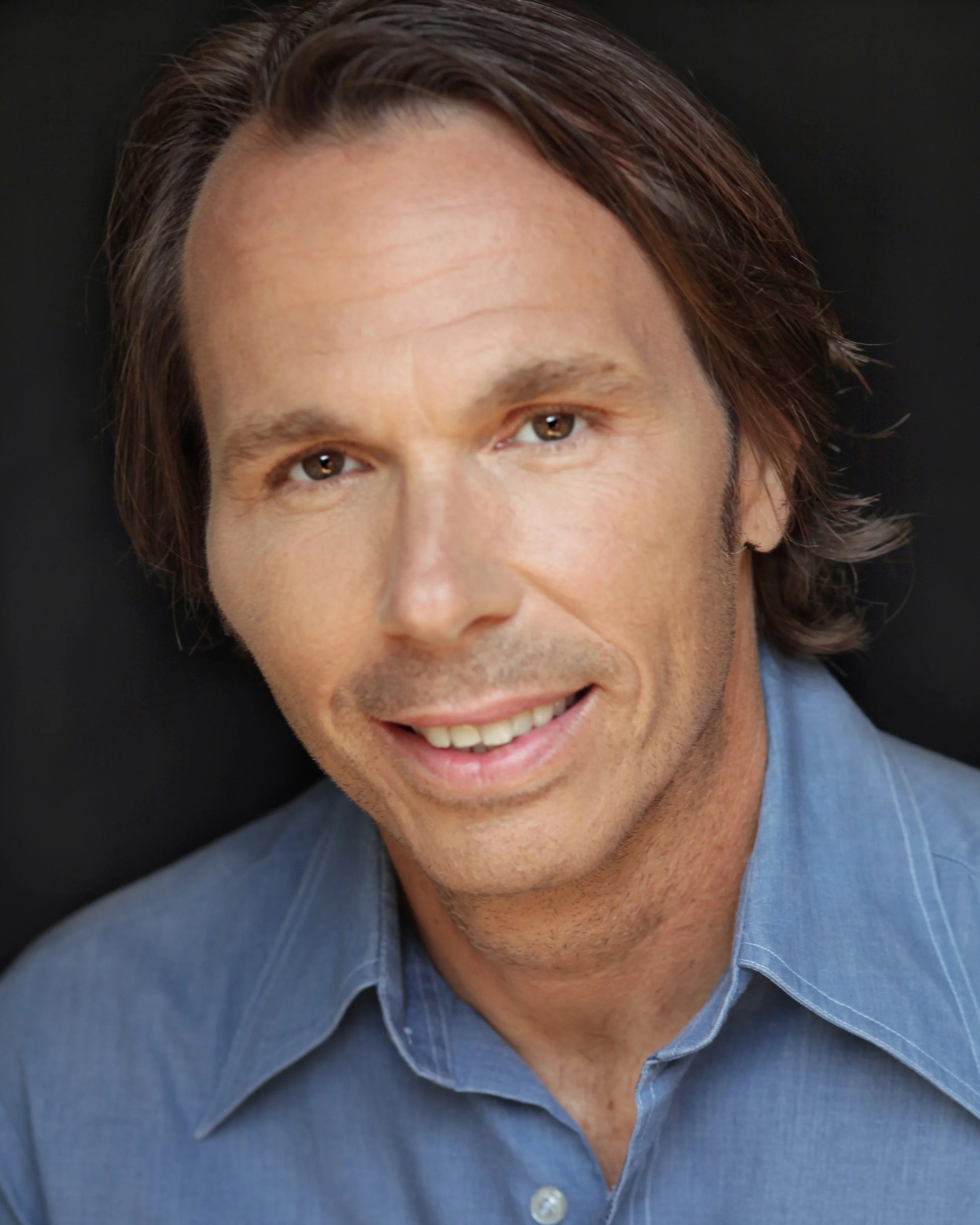 Steven O'Brien, co-host of the Steve and Nadia Save the World Podcast. Close-up of a man's face, He has brown eyes, light skin, and medium-length brown hair. He is wearing a blue shirt with the top button undone. The background is black.