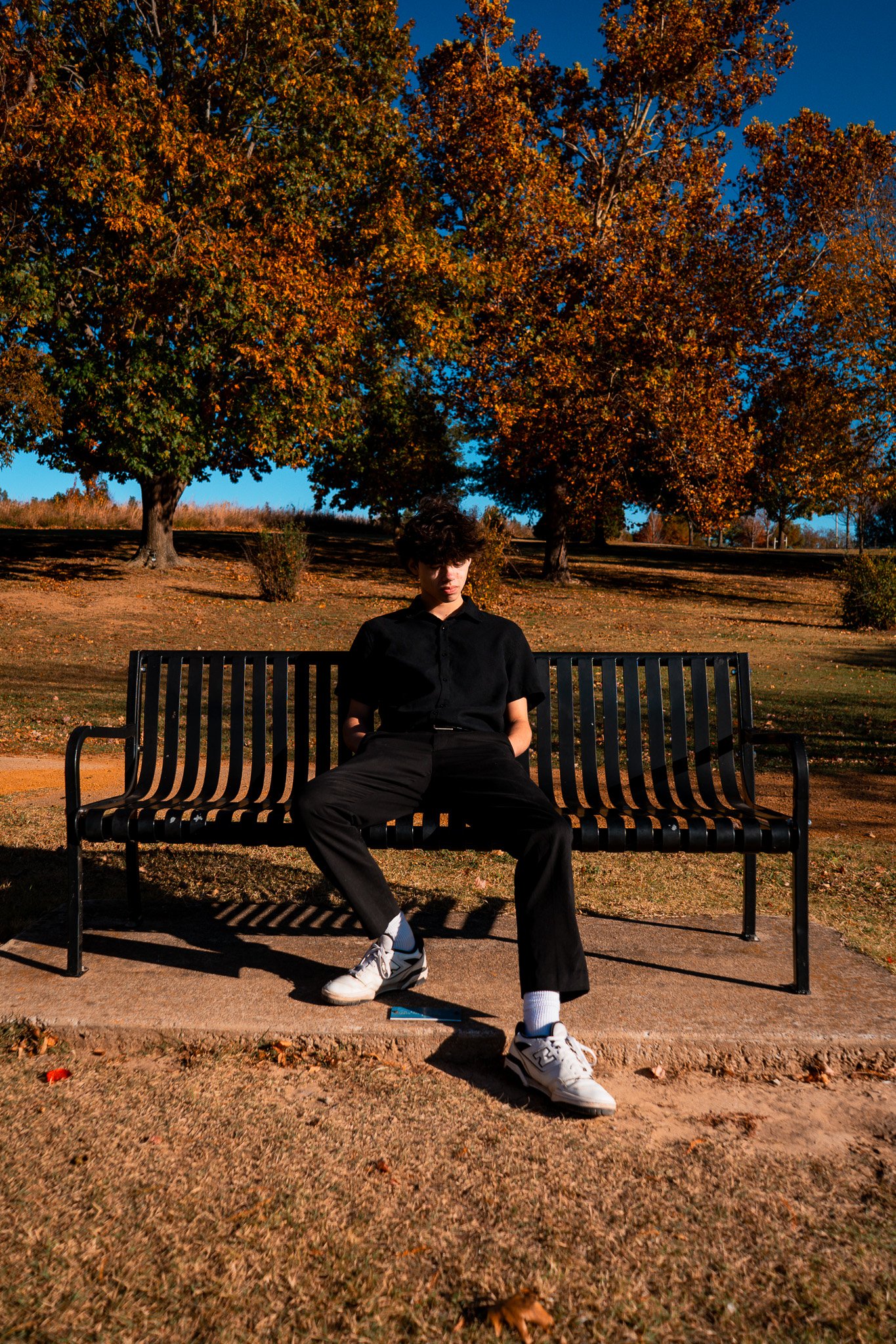 A young man with dark, wavy hair is sitting on a black metal park bench outdoors during autumn. He is wearing a black shirt, black pants, white sneakers, and white socks. The park has large trees with orange, yellow, and green leaves, and the ground is covered with fallen leaves. The sky is bright blue with no clouds visible.