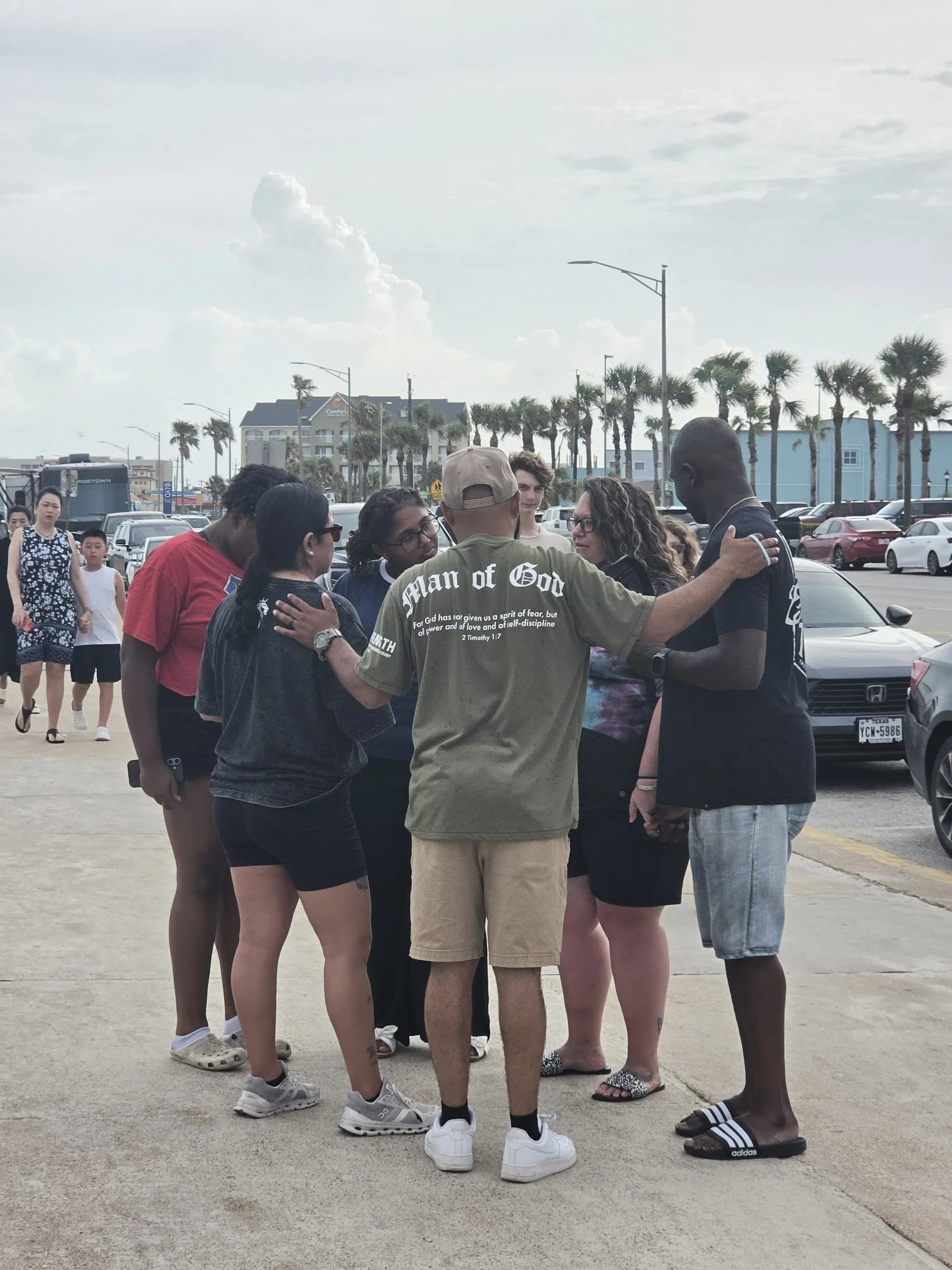 A diverse group of people standing in a circle outdoors, holding hands and praying. They are in a parking lot with cars, palm trees, and buildings in the background on a partly cloudy day.