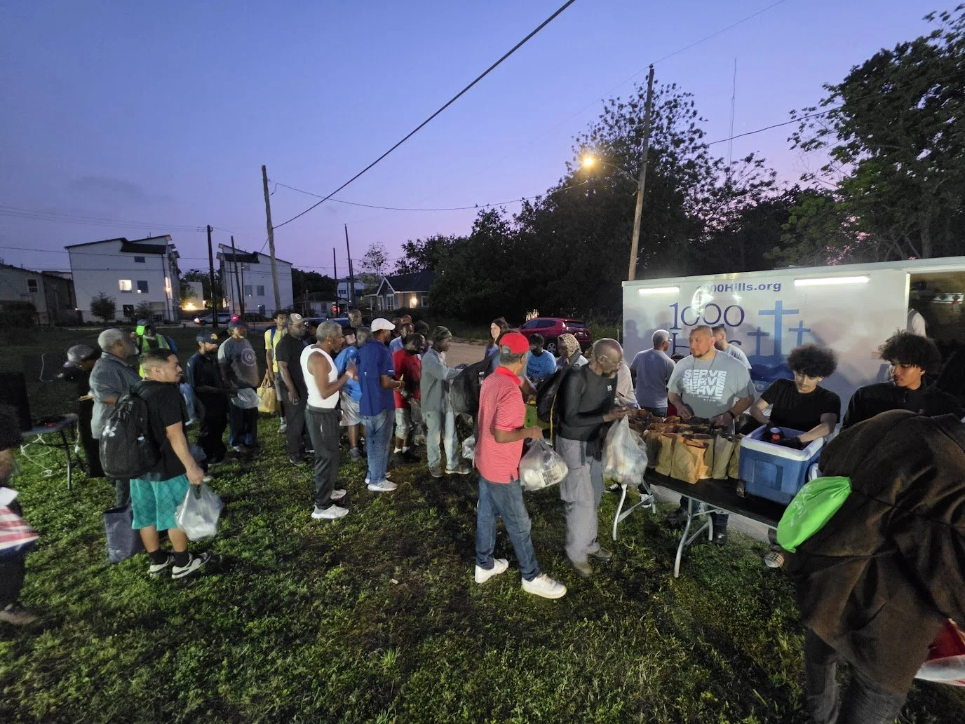 A line of people waiting outside for food distribution at dusk, with volunteers handing out packages and a truck in the background with a church logo and website.