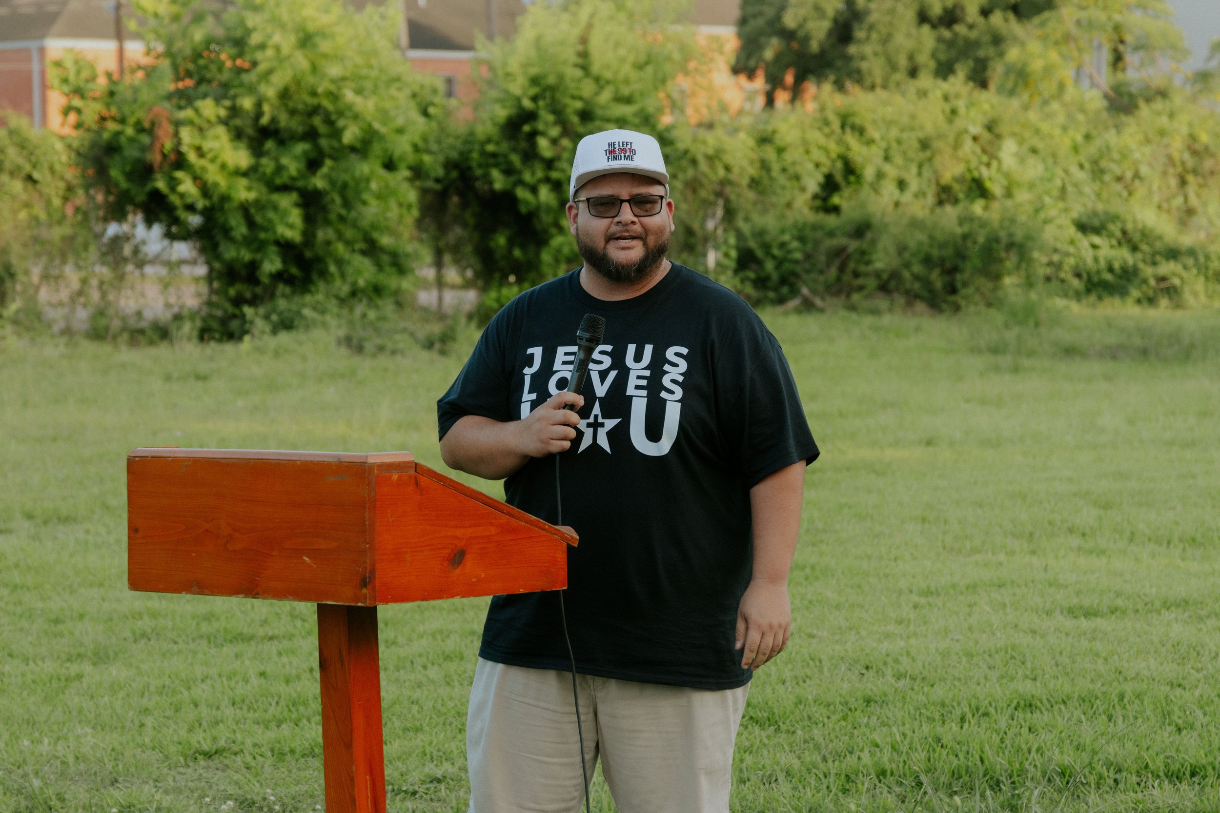 A man with glasses and a beard, wearing a white baseball cap and a black T-shirt with religious text, stands outdoors in front of a green grassy area and trees. He is holding a microphone near a wooden podium.