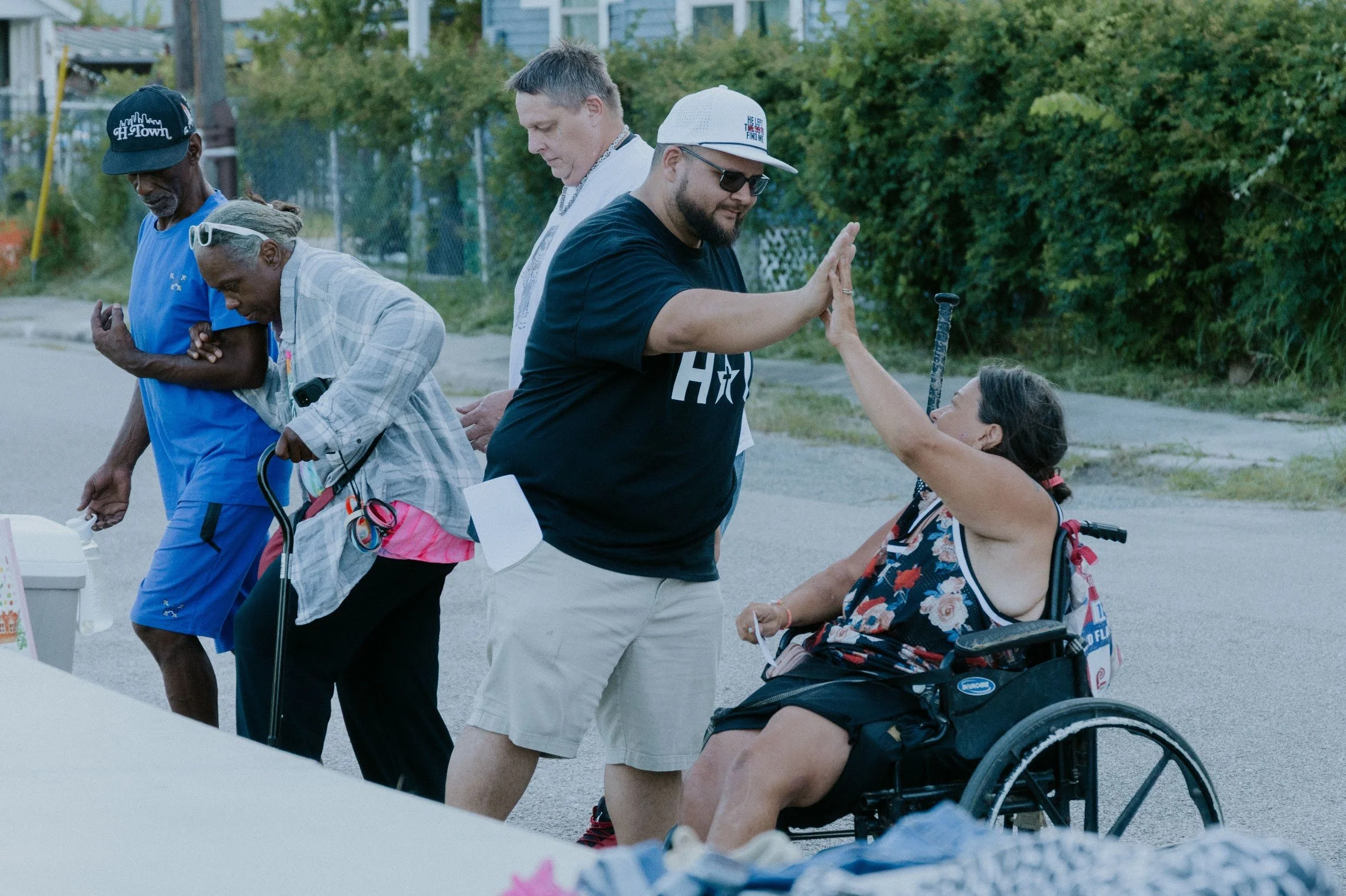 Group of five people, including a woman in a wheelchair, participating in a high five during an outdoor event in a park with trees in the background.