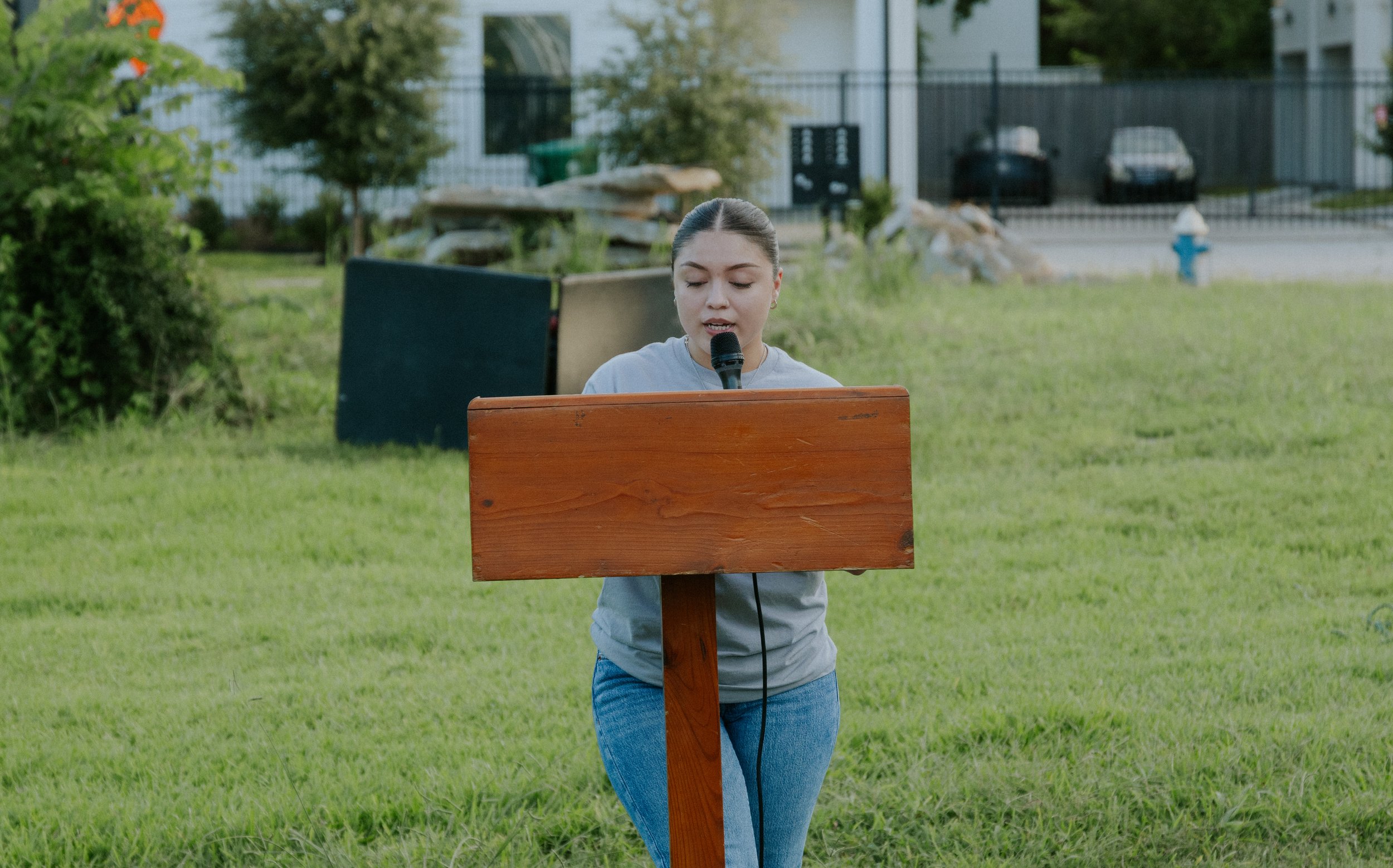 Young woman speaking into a microphone at a wooden podium outdoors on grassy area.
