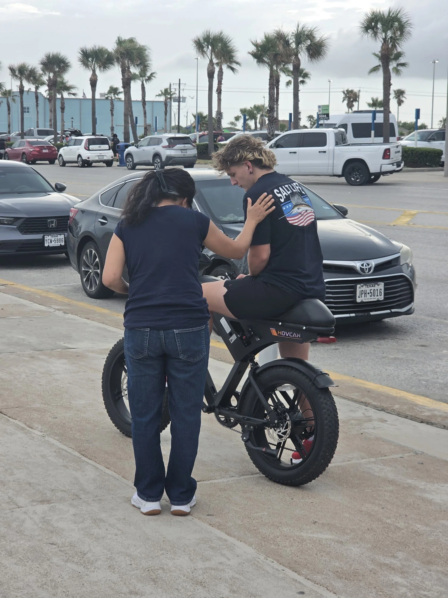 Two people, one with long dark hair and the other with short blond hair, are sitting on a black electric bike in a parking lot with cars and palm trees in the background. The person with blond hair is wearing a shirt with an American flag design and 