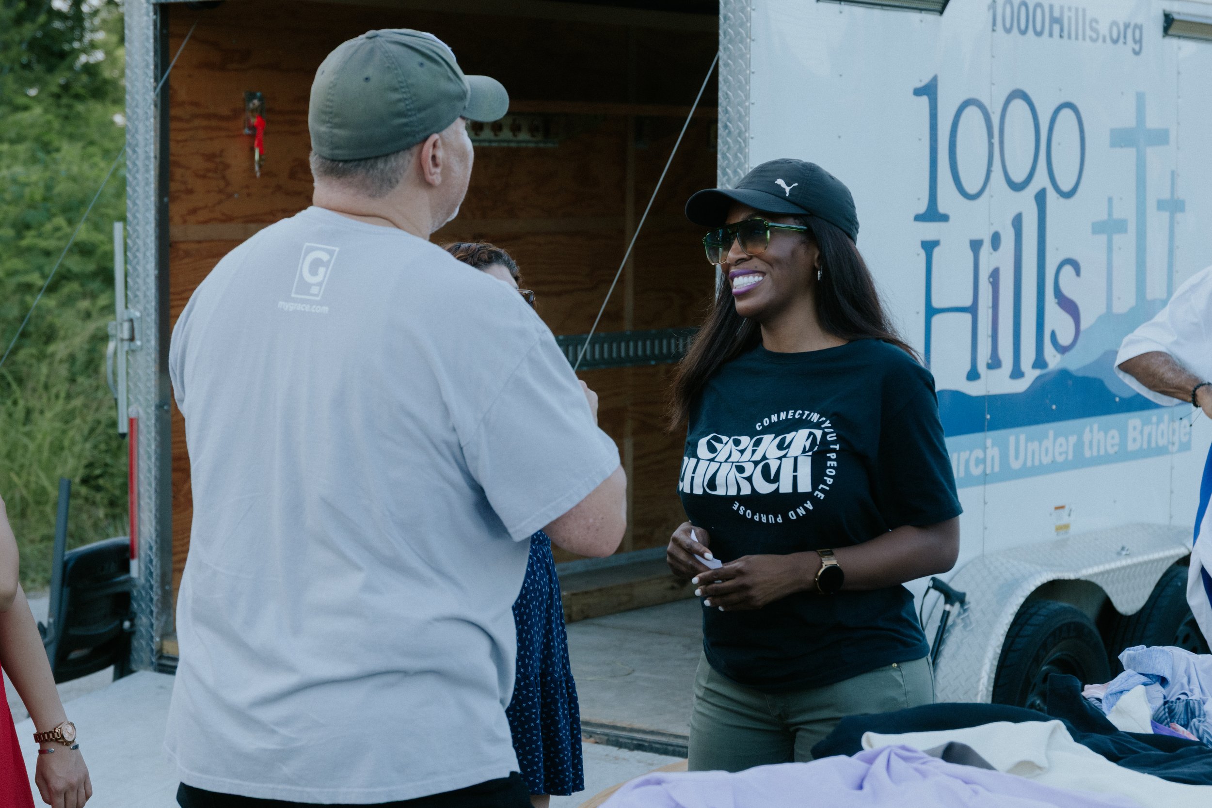 Two women are standing outside and talking. One woman has darker skin, black hair, sunglasses, a black cap, and a black T-shirt. The other woman has lighter skin, short hair, and is wearing a light-colored shirt and gray cap. They are near a mobile b