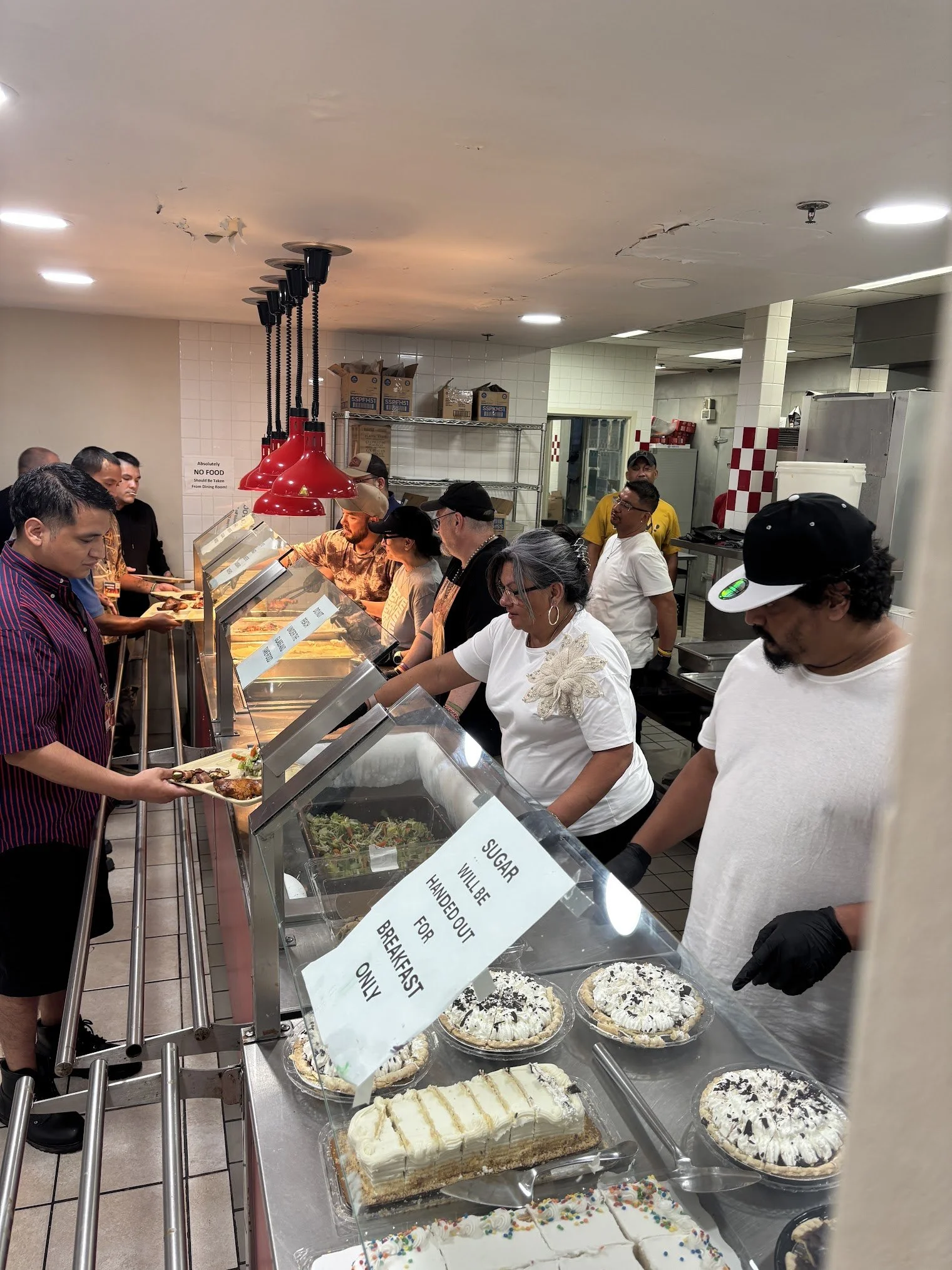 People standing in line at a cafeteria-style food service counter with desserts like cakes, pies, and ice cream in display cases.