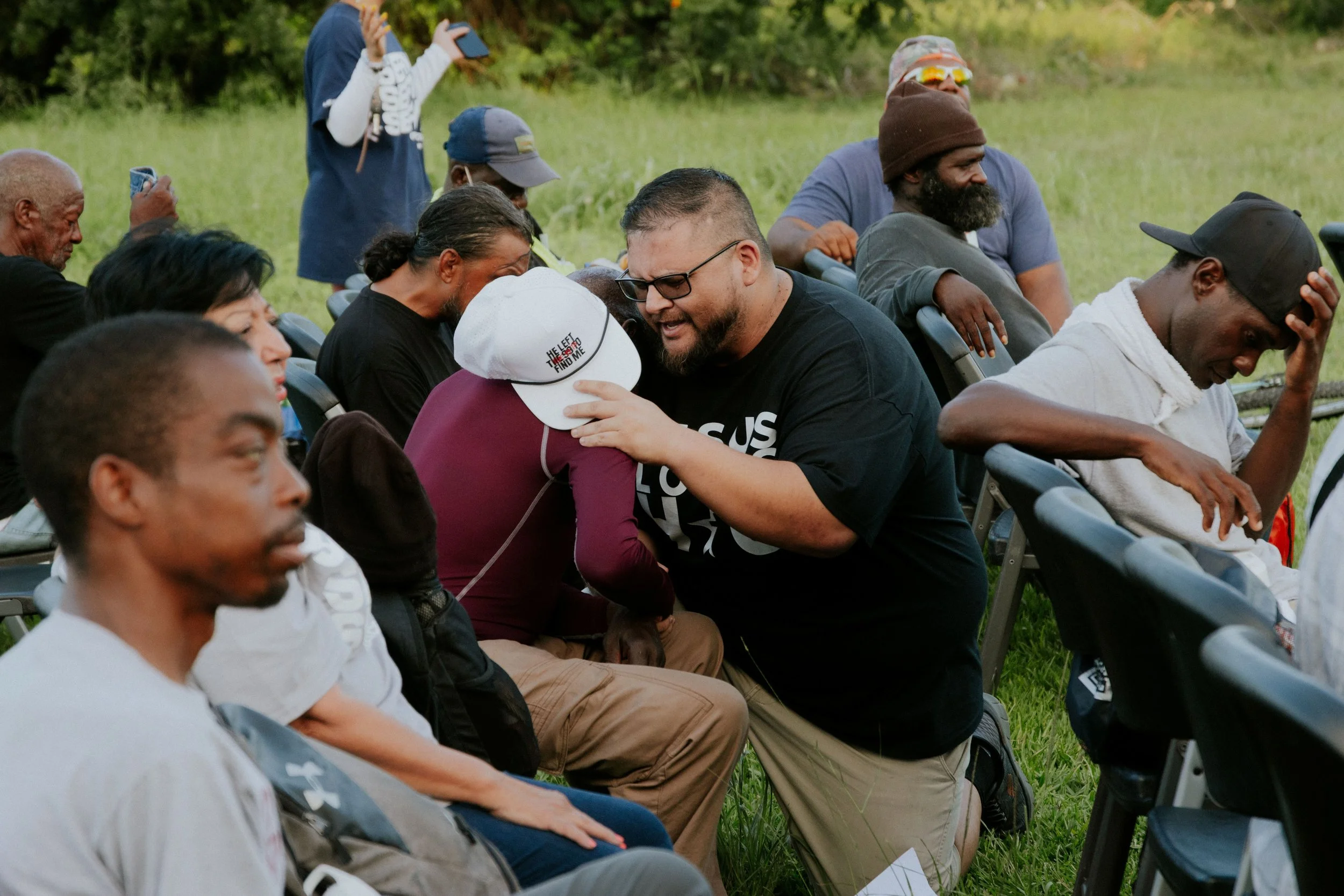 Group of people seated outdoors on chairs in a grassy area, some with eyes closed, one person praying, others looking contemplative, and one individual showing a gesture of blessing.