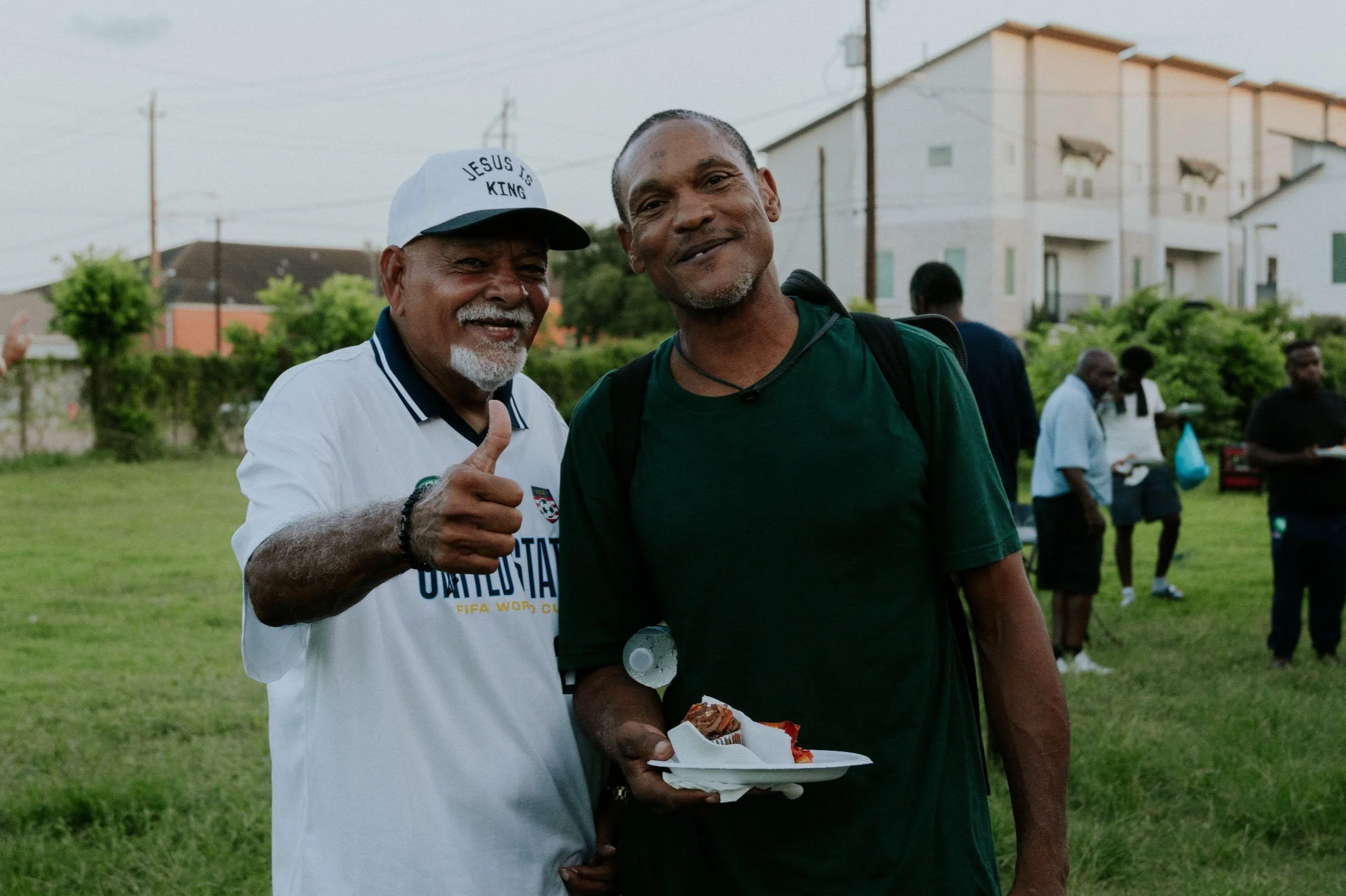 Two men standing outdoors on a grassy area, smiling at the camera. The man on the left is wearing a white cap with the words "JESUS IS KING" and a white shirt with text, giving a thumbs-up. The man on the right is wearing a green shirt and has a back