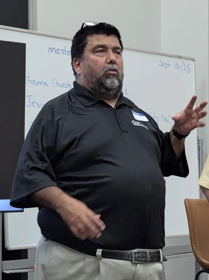 A man with dark hair, beard, and glasses on his head, wearing a black polo shirt, appears to be speaking or explaining something in front of a whiteboard in a meeting or classroom setting.