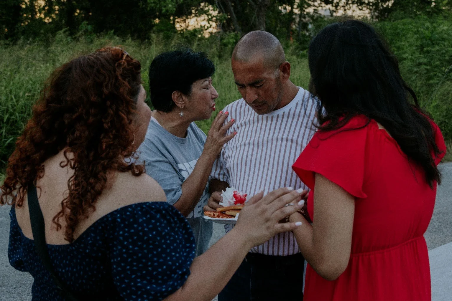 Four women and one man standing together outdoors, with one woman in a red dress holding a slice of cake with red icing, appearing emotional and comforting the man, who is holding a plate of cake and wearing a striped t-shirt.