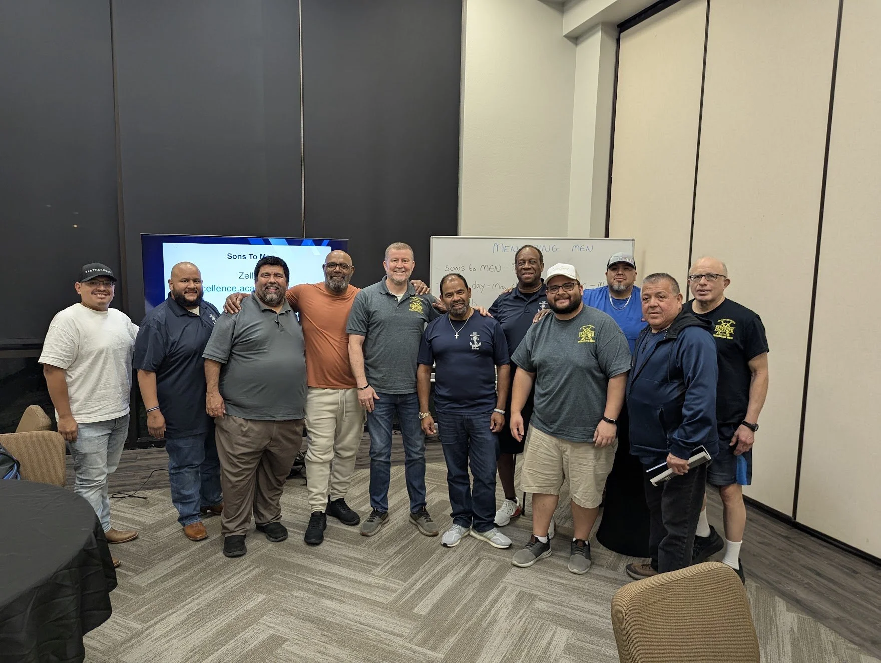 A group of twelve men standing together in a conference room, smiling for a photo. Behind them, a screen displays a presentation slide with the title 'Sons To Men' and a whiteboard shows notes related to mentoring men.