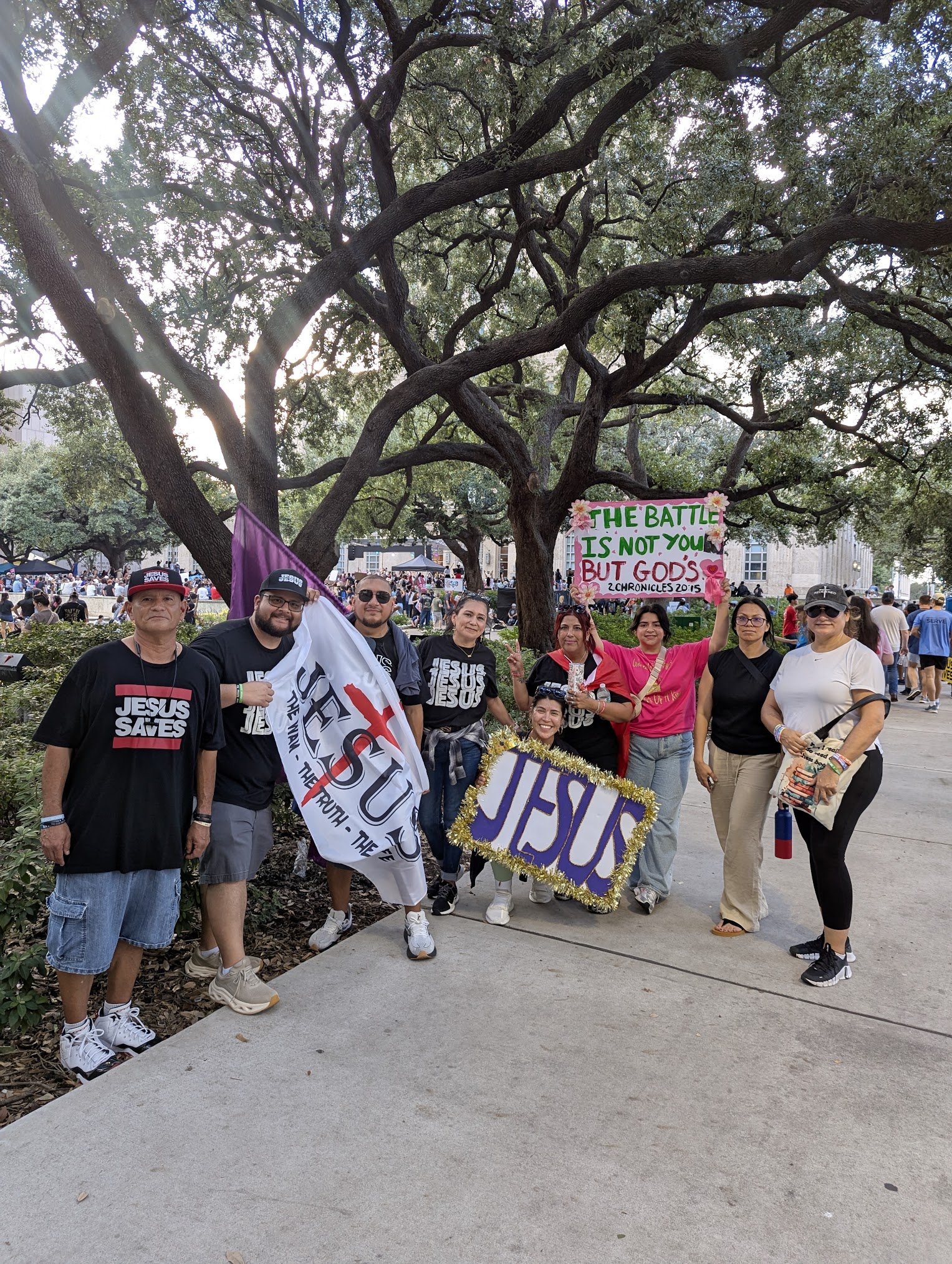 A group of people gathered outdoors in a park, holding signs and flags during a demonstration or rally, with large trees and a crowd in the background.